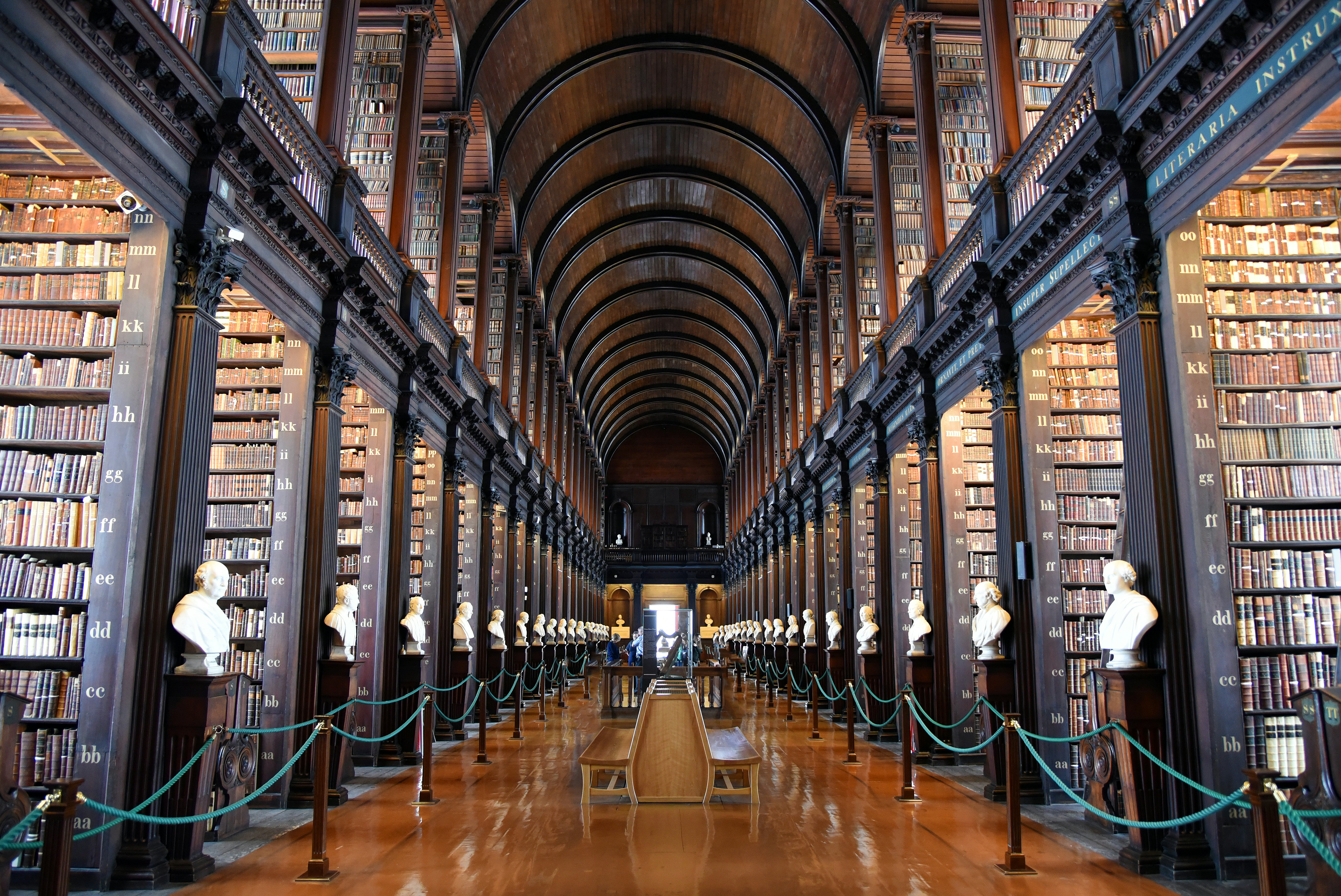 Two stories of floor-to-ceiling bookcases are lined up on either side of a central walkway with a wood floor and benches in the center; there are white busts at the end of each bookcase.