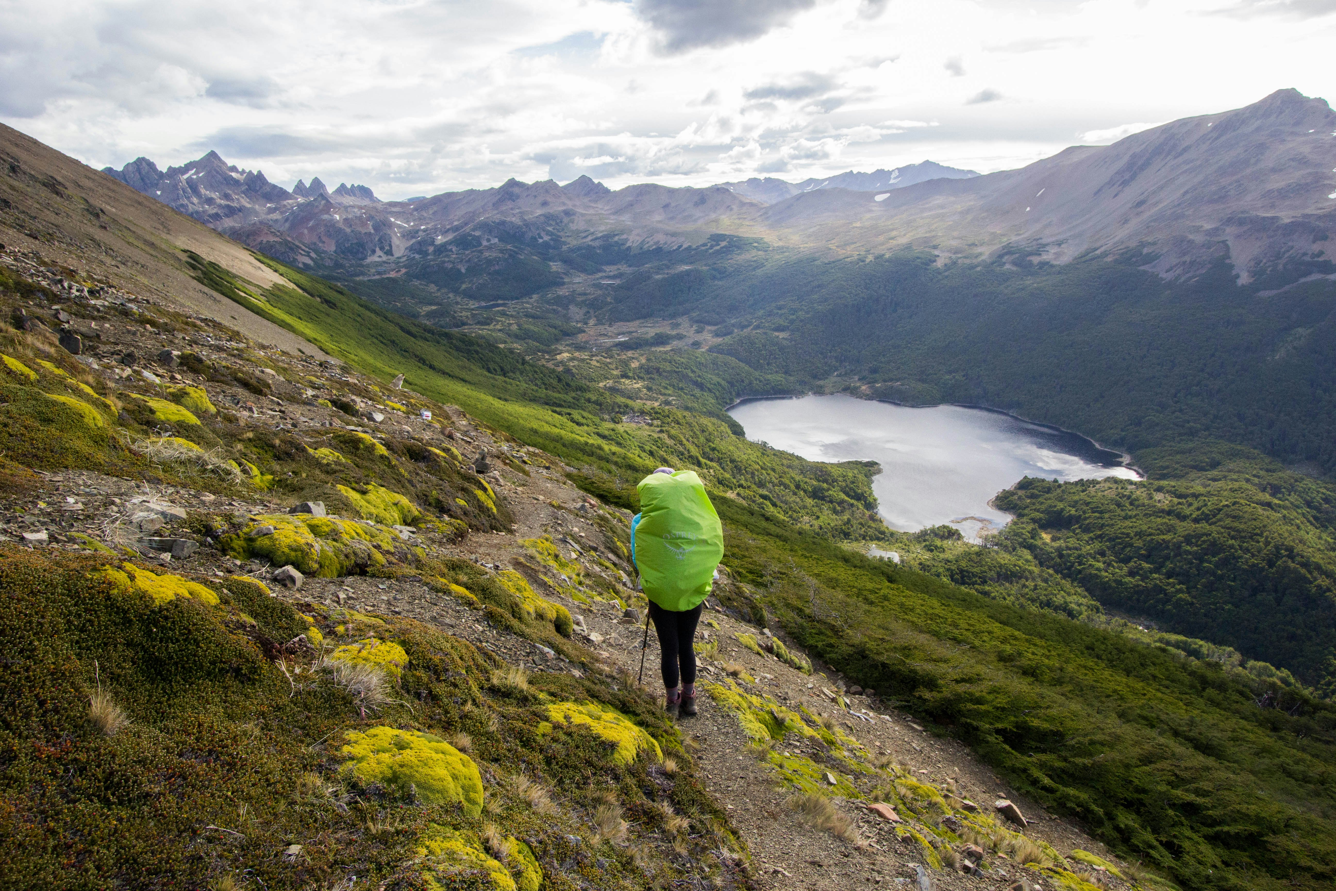 A hiker with a big covered backpack navigates a hillside in a mountainous landscape. A lake and mountains are seen in the distance.