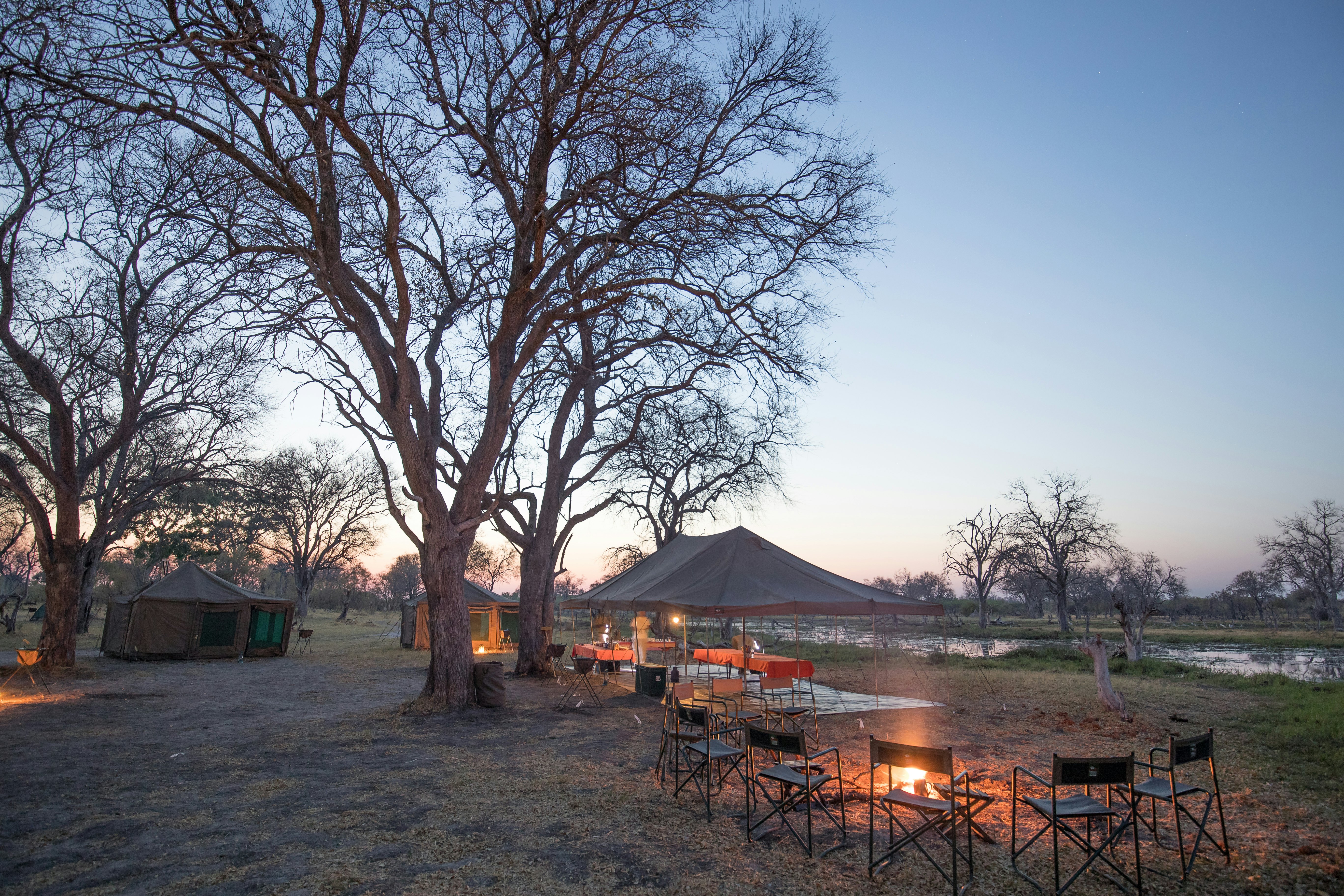 Camp chairs around a fire and a large canopy set up at dusk at a campsite near a river.