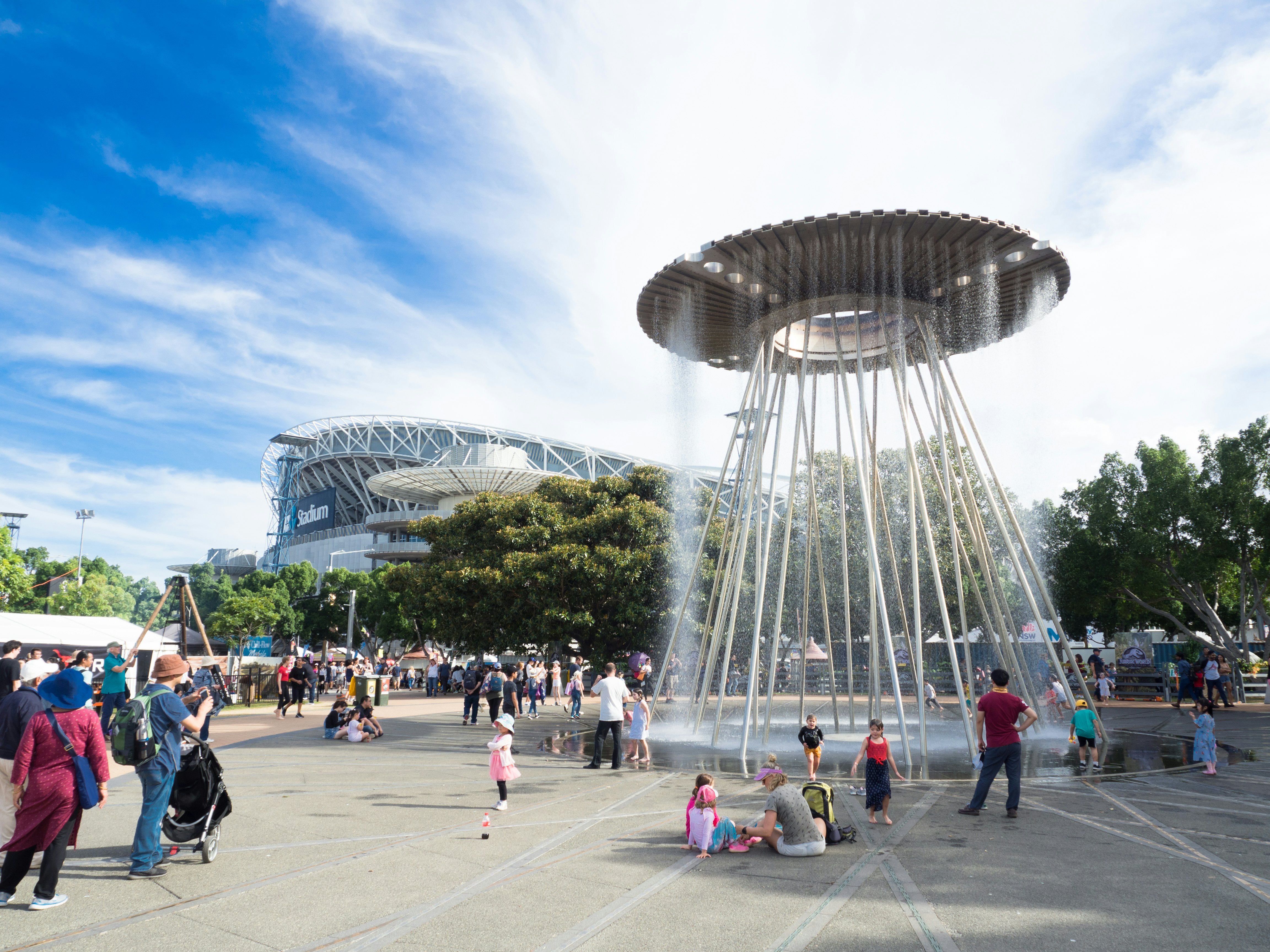 Families gather around a large fountain with circular top in the middle of a public sqaure on a sunny day.