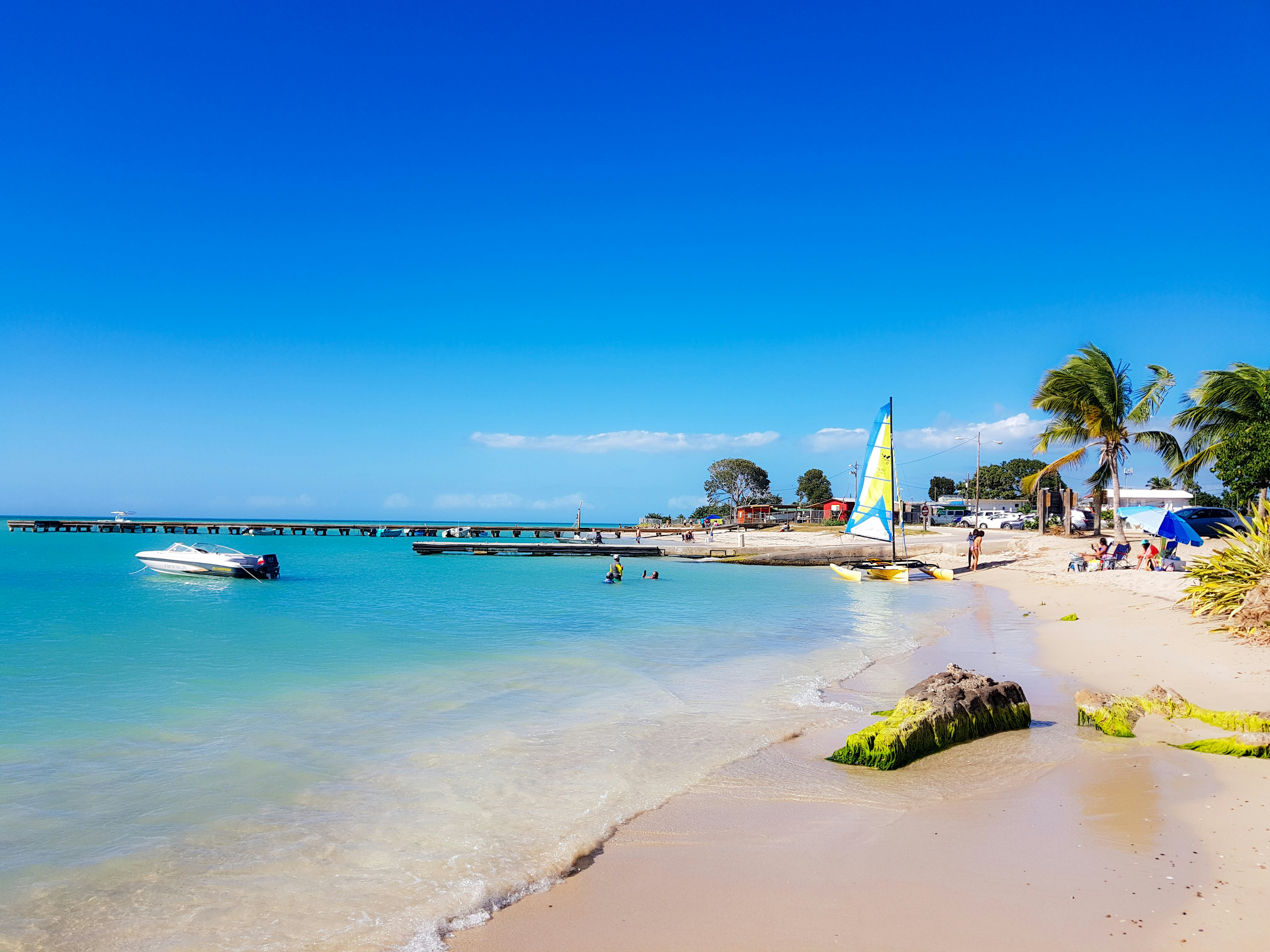 A sandy beach with turquoise ocean to the left and bright blue sky above. A catamaran and a few people are on the beach in the distance. A power boat is moored close off shore, and a long pier juts into the water in the far distance.