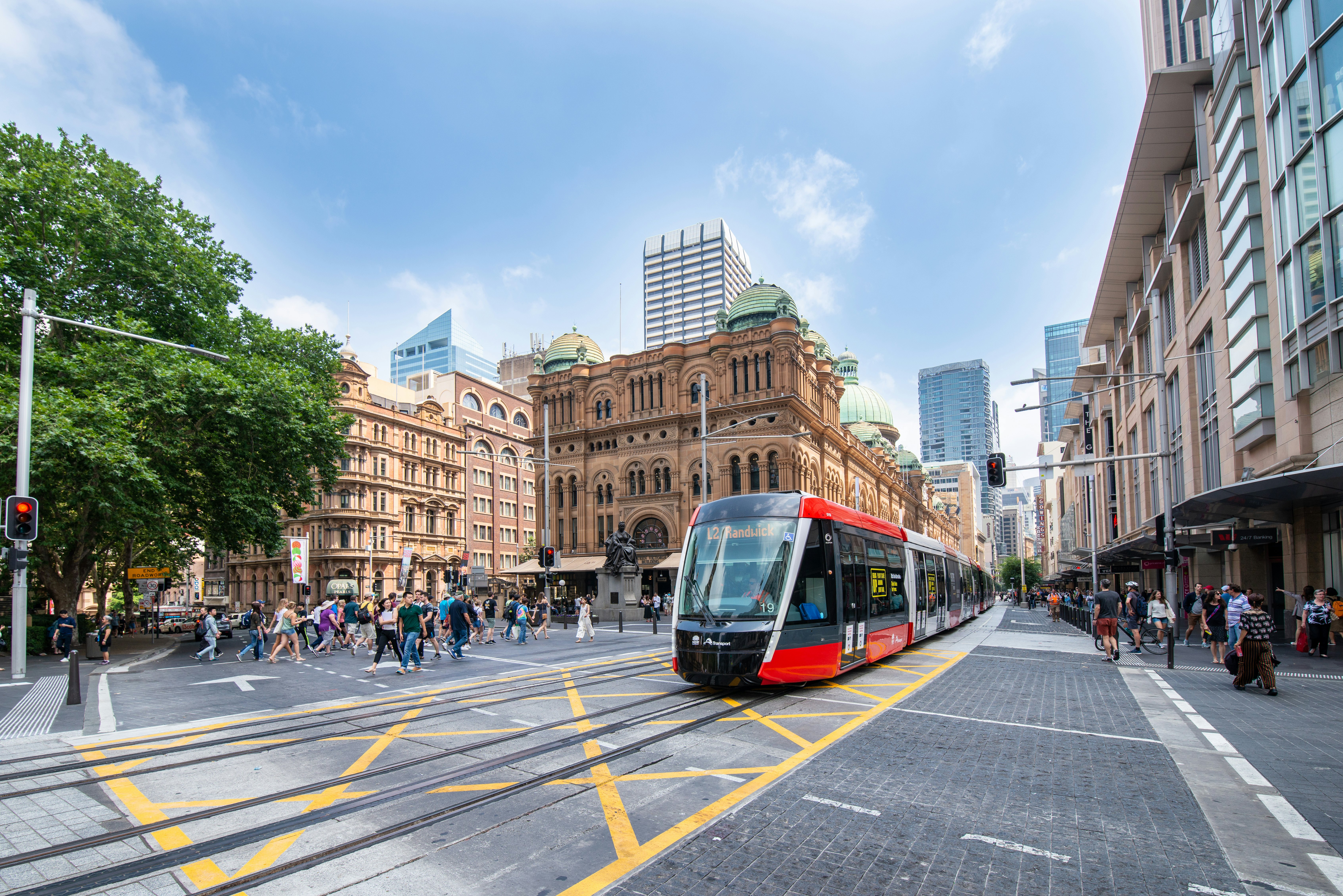A red, modern tram on tracks traveling through a city square on a sunny day. Pedestrians walk on the sidewalks on either side of the train.