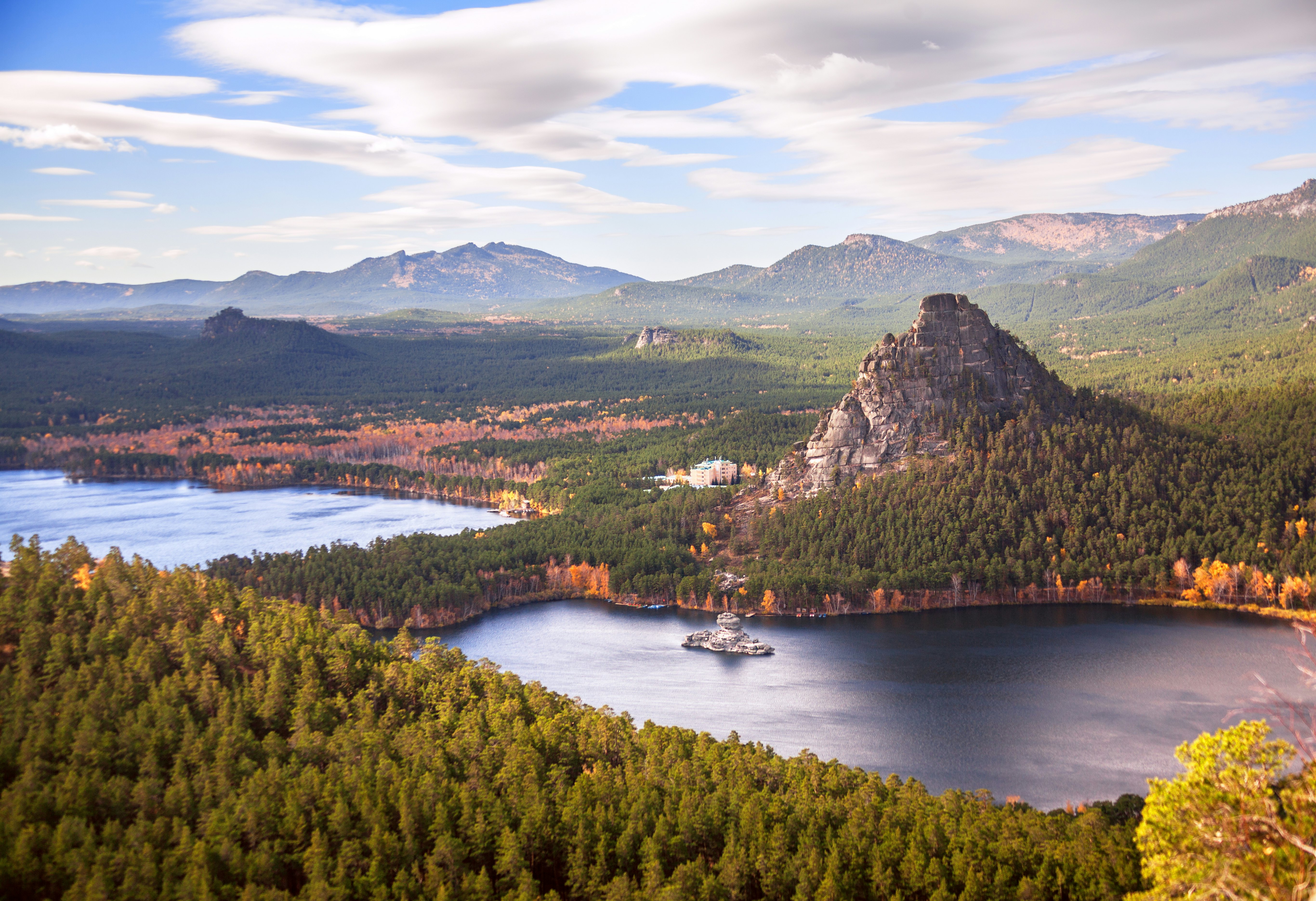 A wide view looking down at a lake surrounded by pine trees, a large rocky peak and other trees showing autumn foliage.