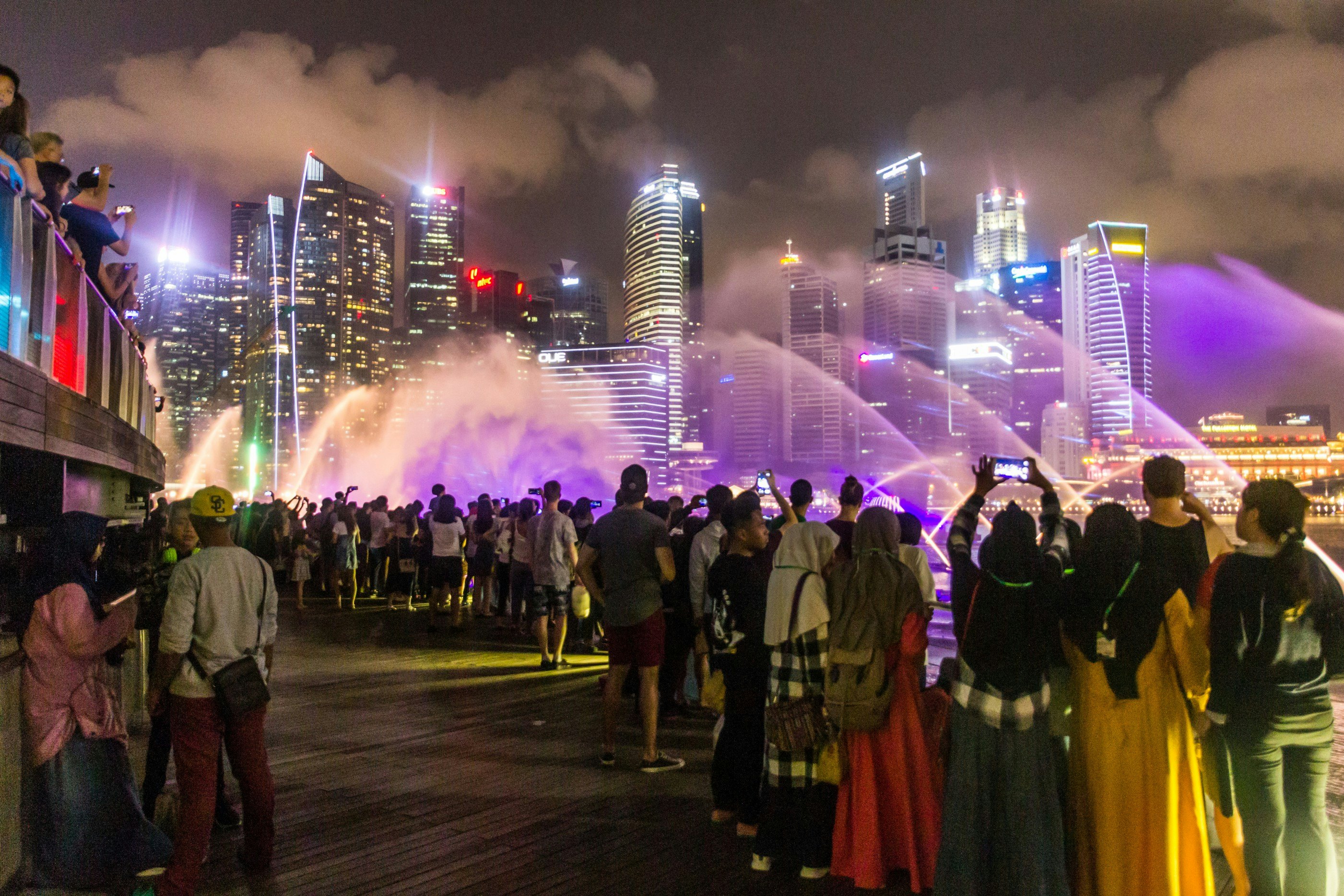 People cluster on an outdoor walkway to watch a presentation of lights and water spray in front of the skyline of a city.