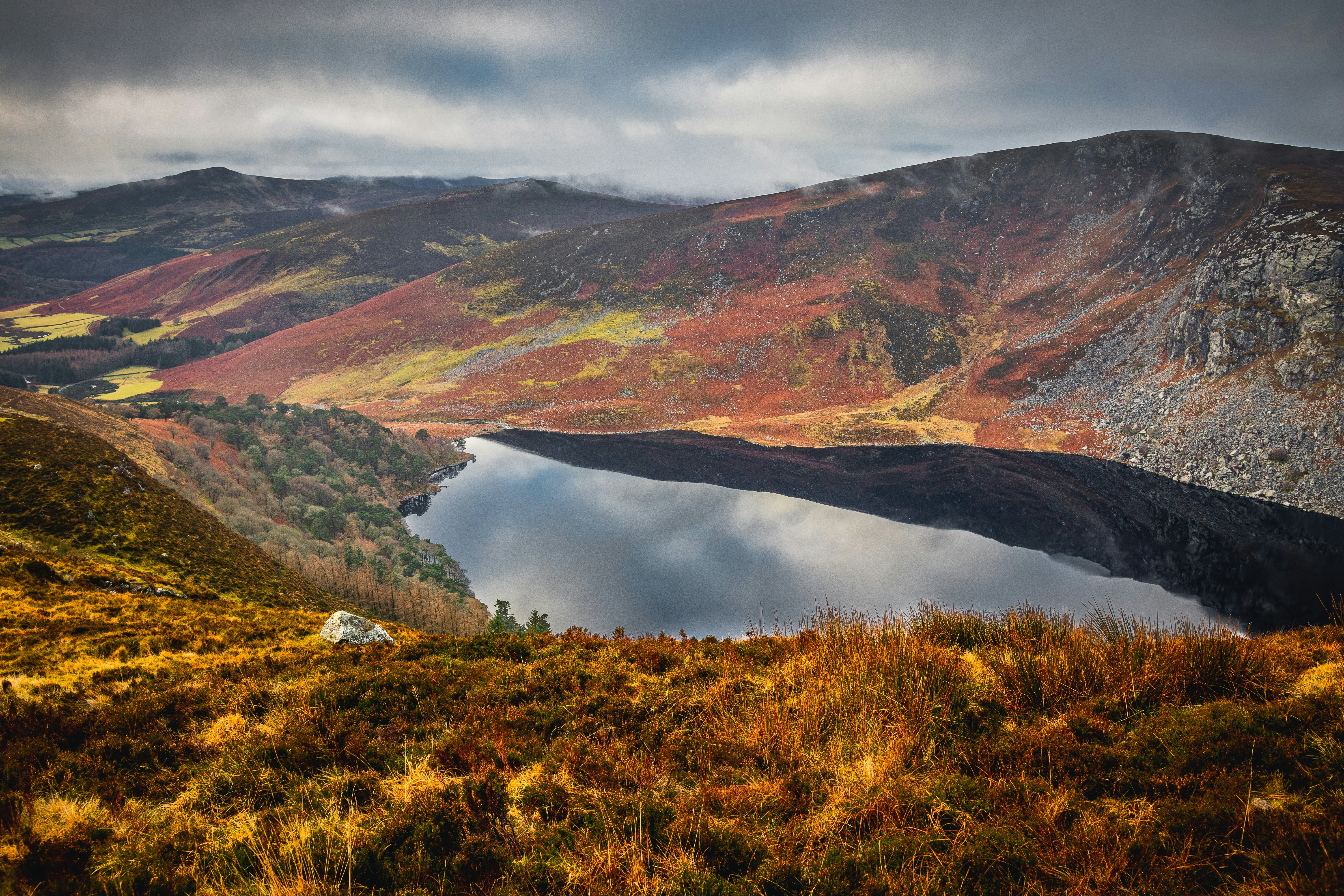 A dark lake surrounded by low hills with fall foliage of yellows and deep orange on an overcast day.