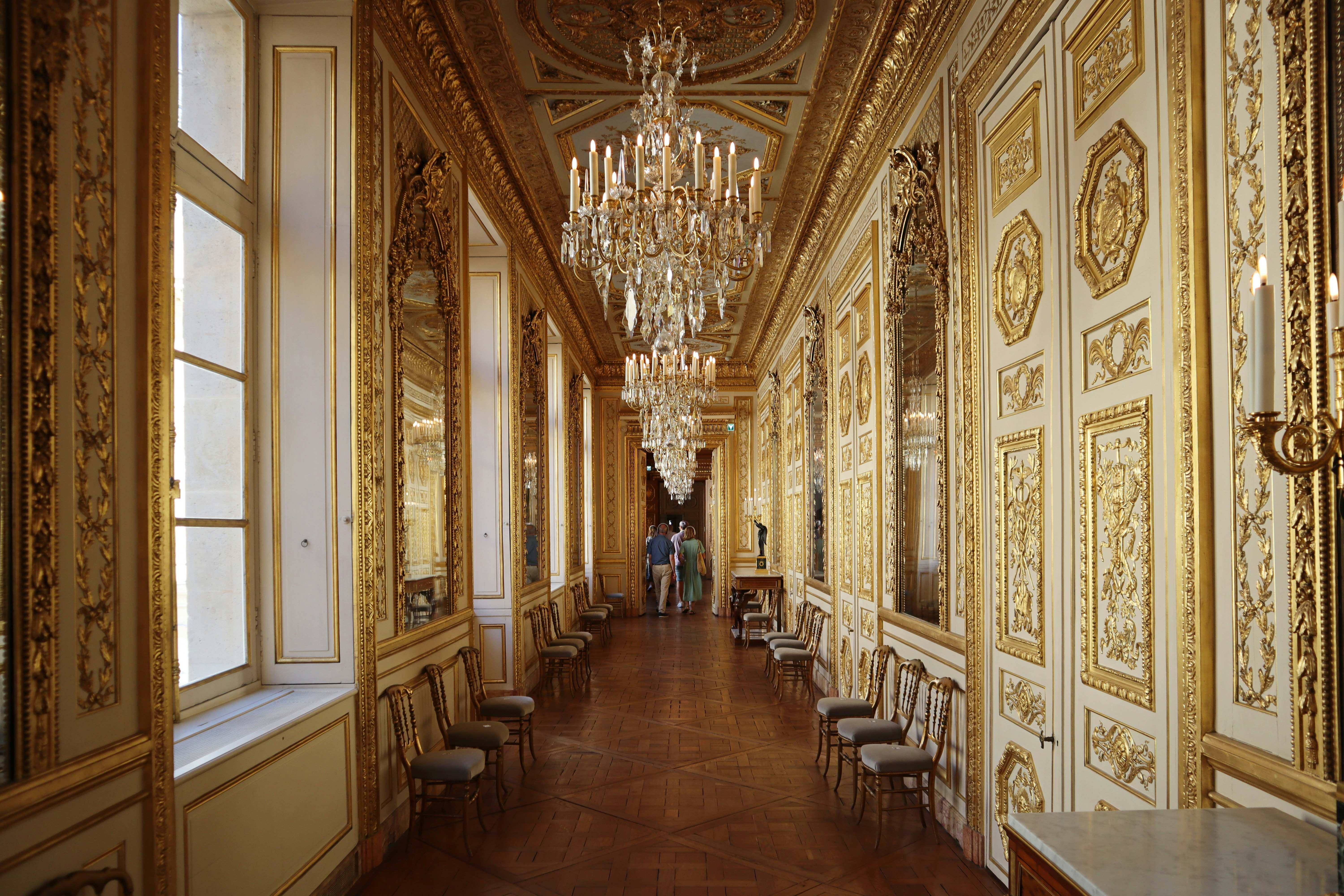A corridor with gold features and details on the woodwork, around the doors and windows. Large chandeliers hang from the ceiling at intervals.