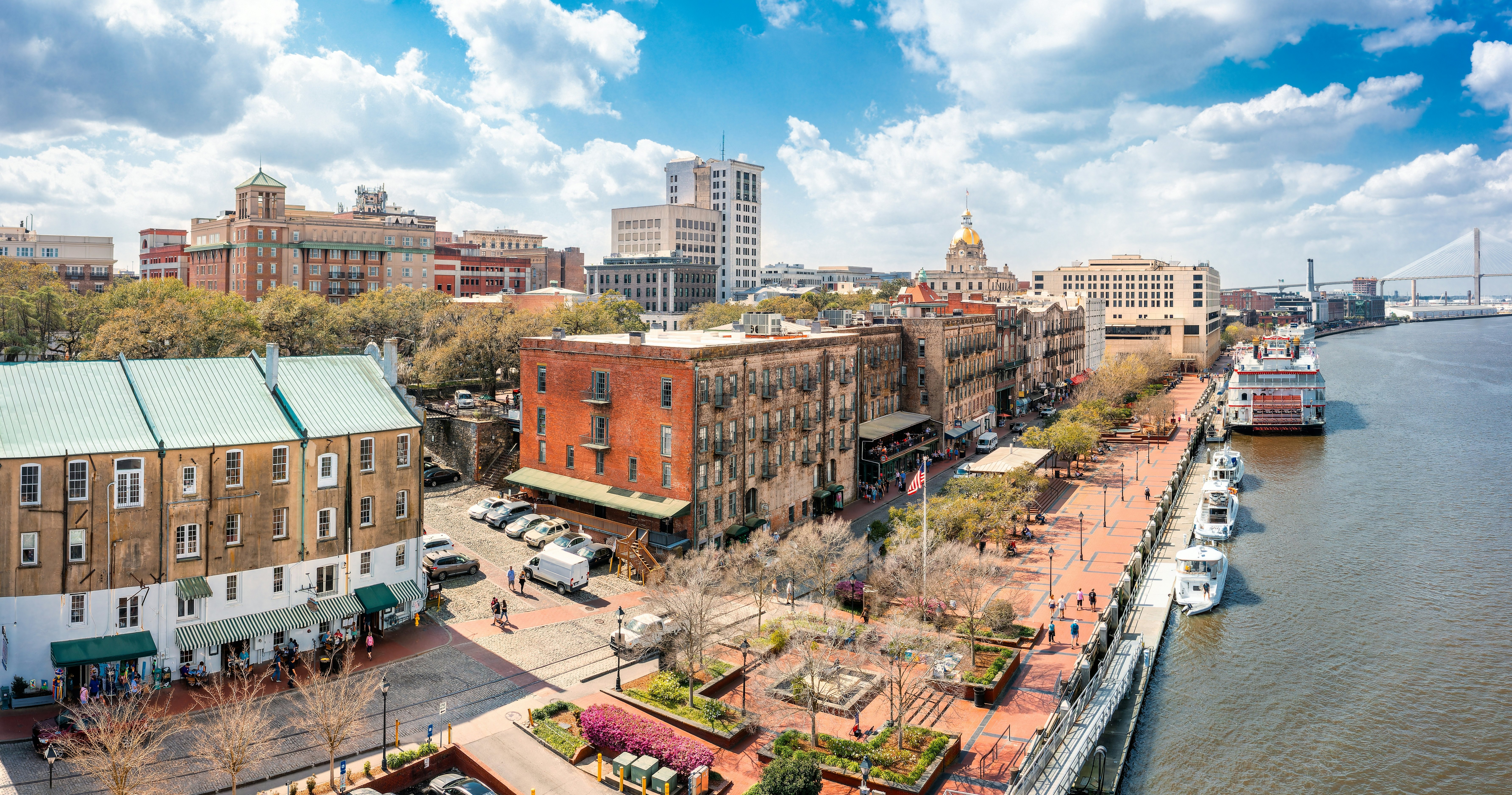 An aerial view of Savannah, Georgia, along the river; a few tall buildings and a building with a gold dome are visible in the distance, as is a suspension bridge.