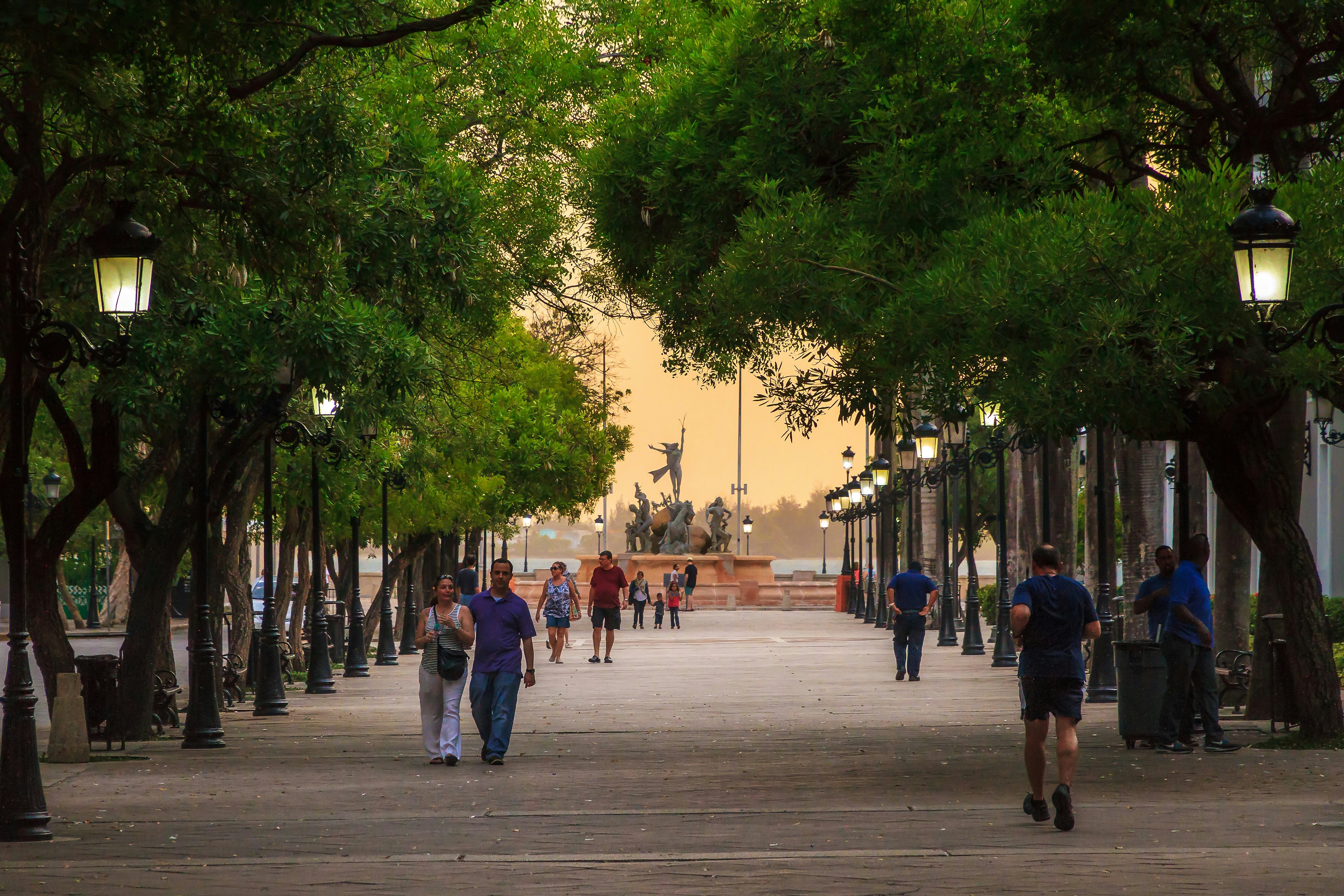 People walk in early evening on a tree-lined pedestrian promenade lit by street lamps in Puerto Rico; a statue is at the end of the path.