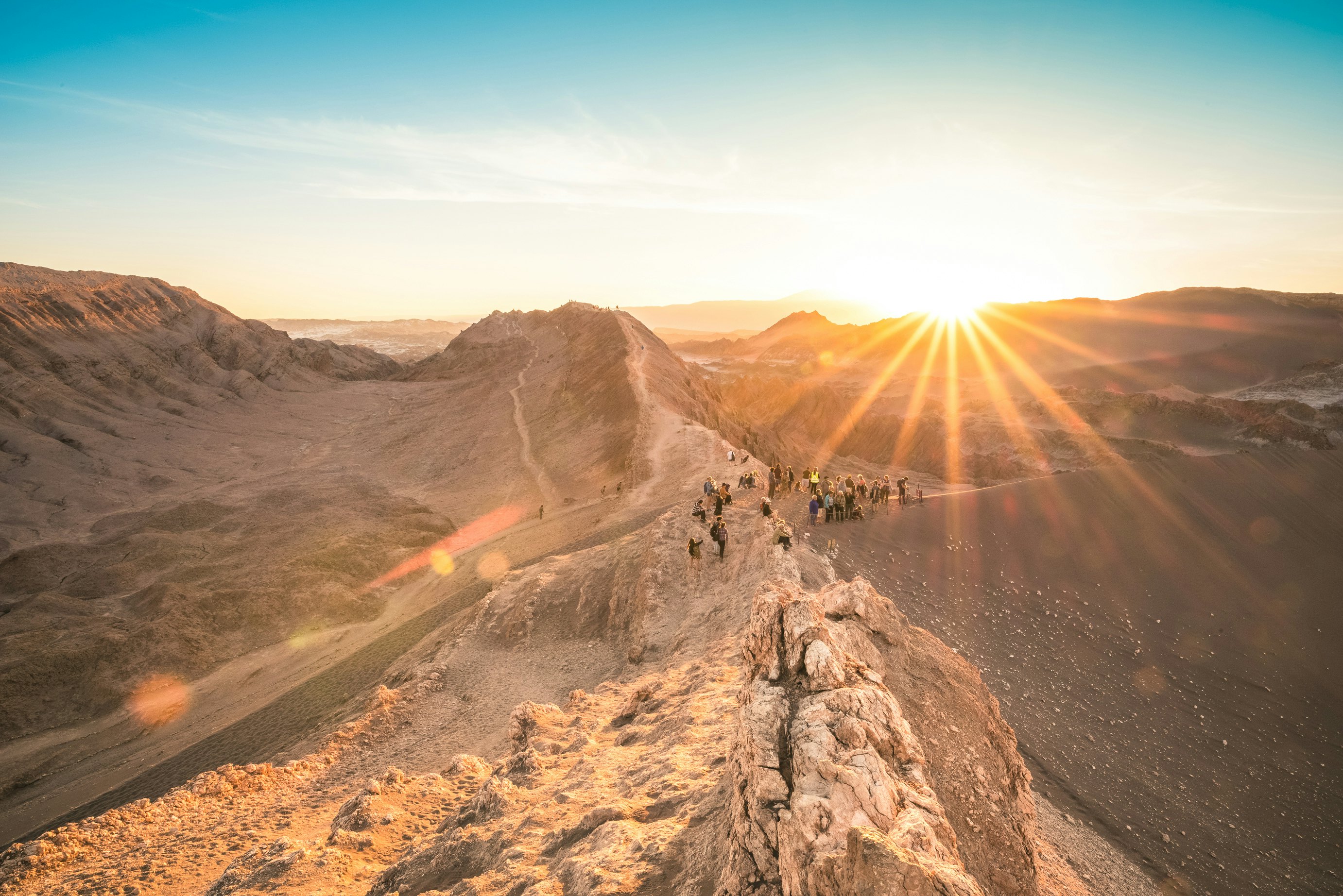 A wide shot of people walking on rock formations in the desert as they watch the sunset.