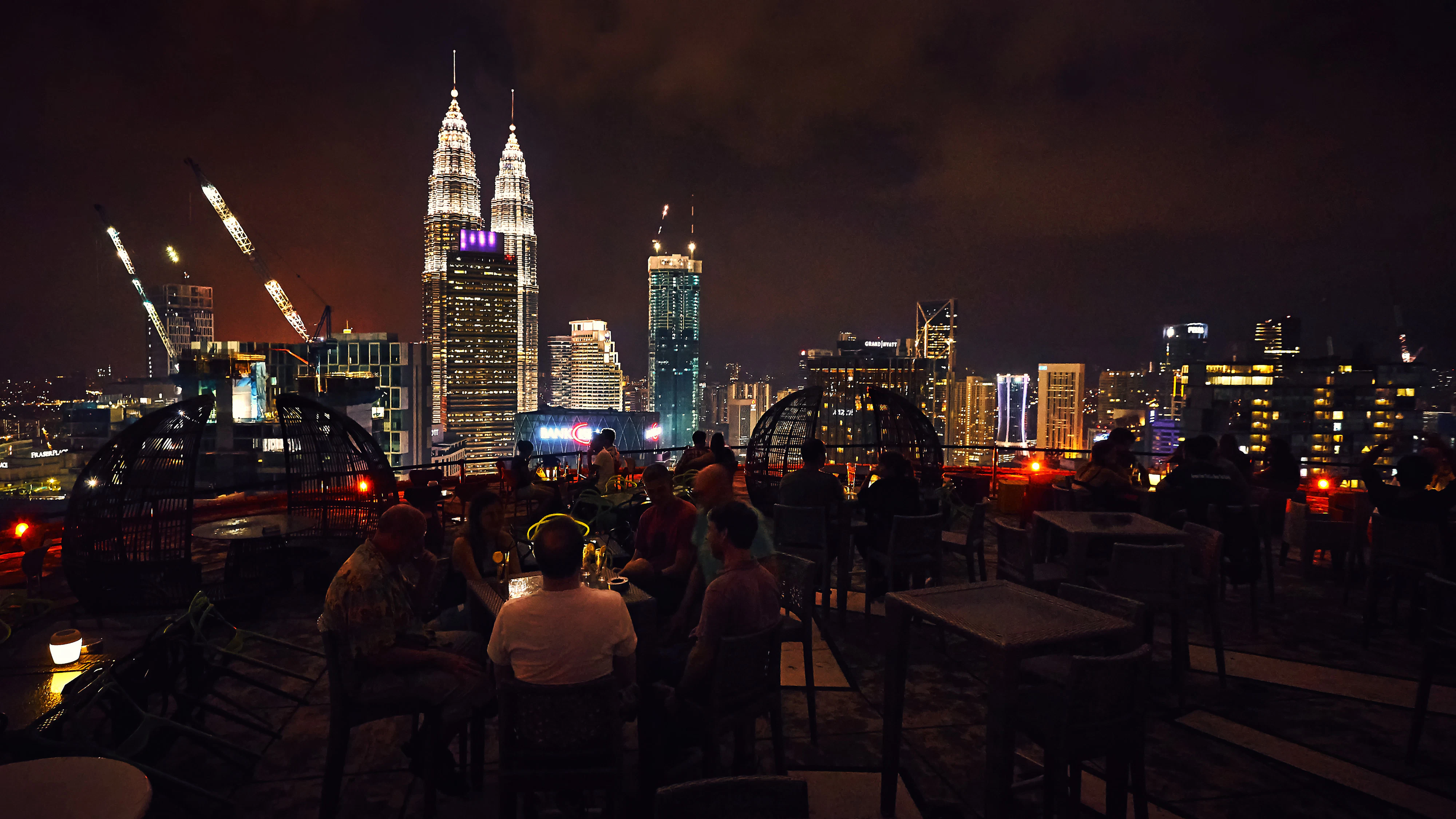 People dine on a rooftop terrace with the skycrapers of a city illuminated in the distance.