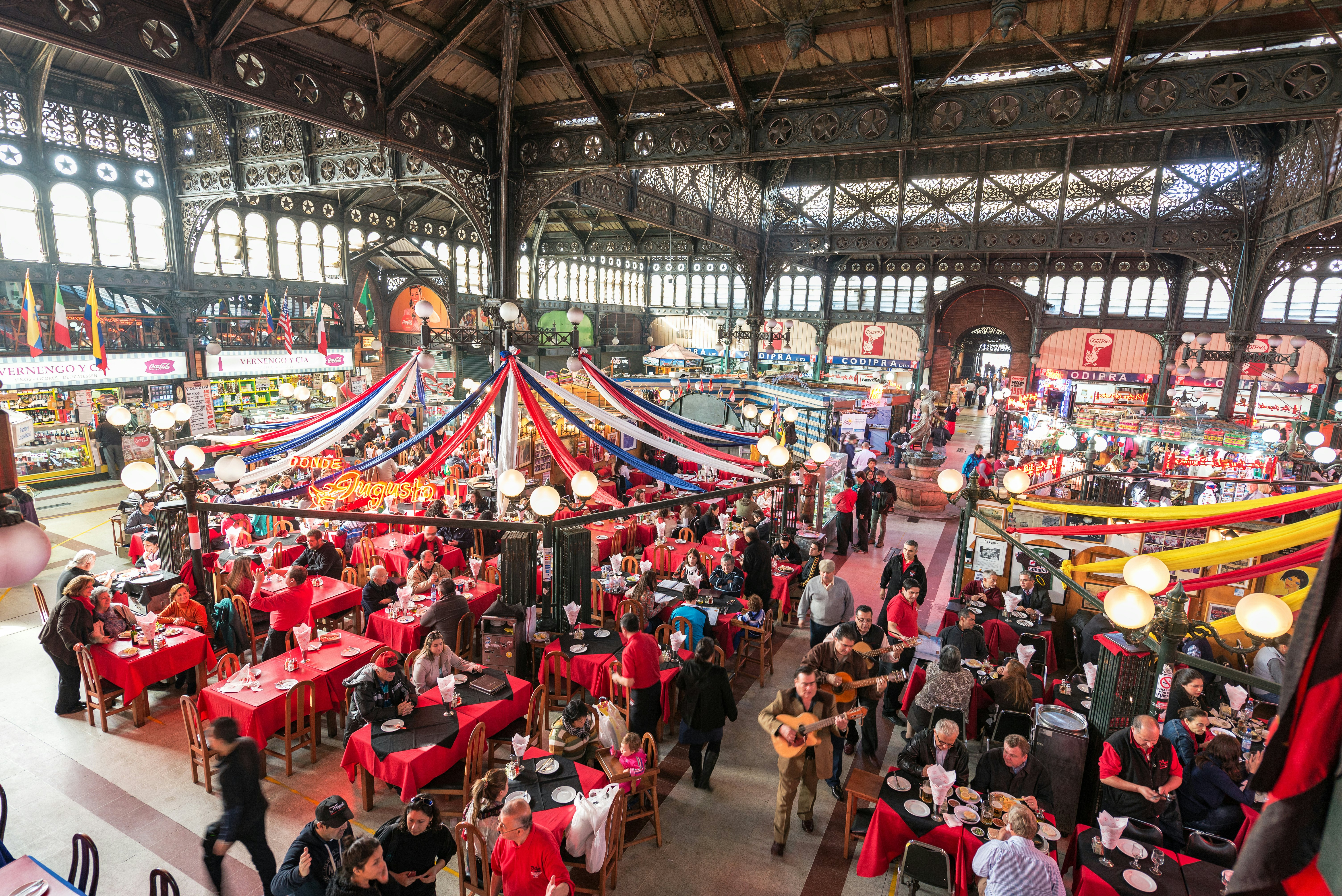 The view of an interior of a market, filled with tables and covered by wrought-iron beams.