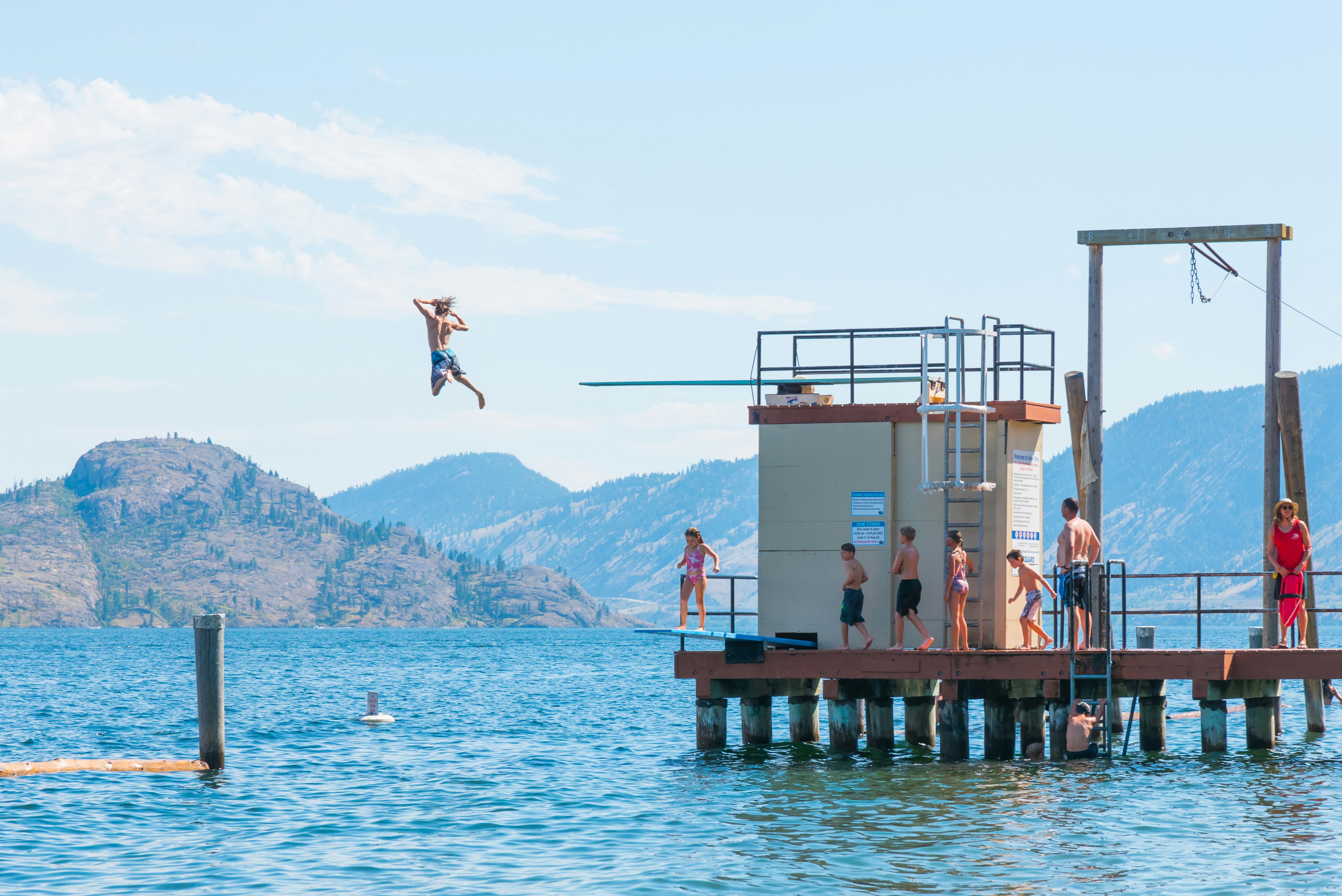 A boy leaps off a dock's diving board into a lake while others stand on the dock watching on a sunny day.