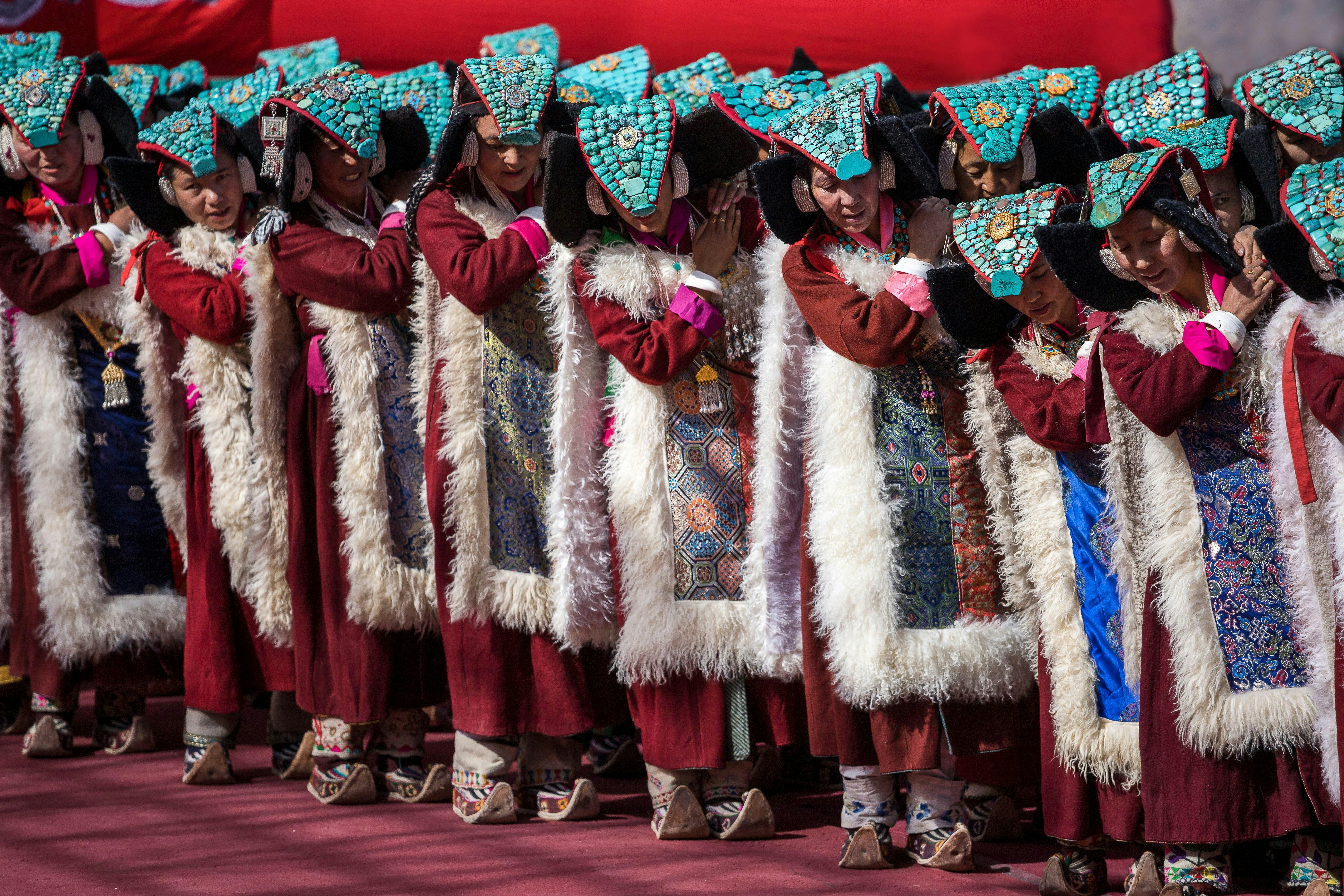 Women in red dresses with fur-lined patterned panels on the front stand in a line as part of a dance formation. Each one has a headdress on embellished with turquoise stones.