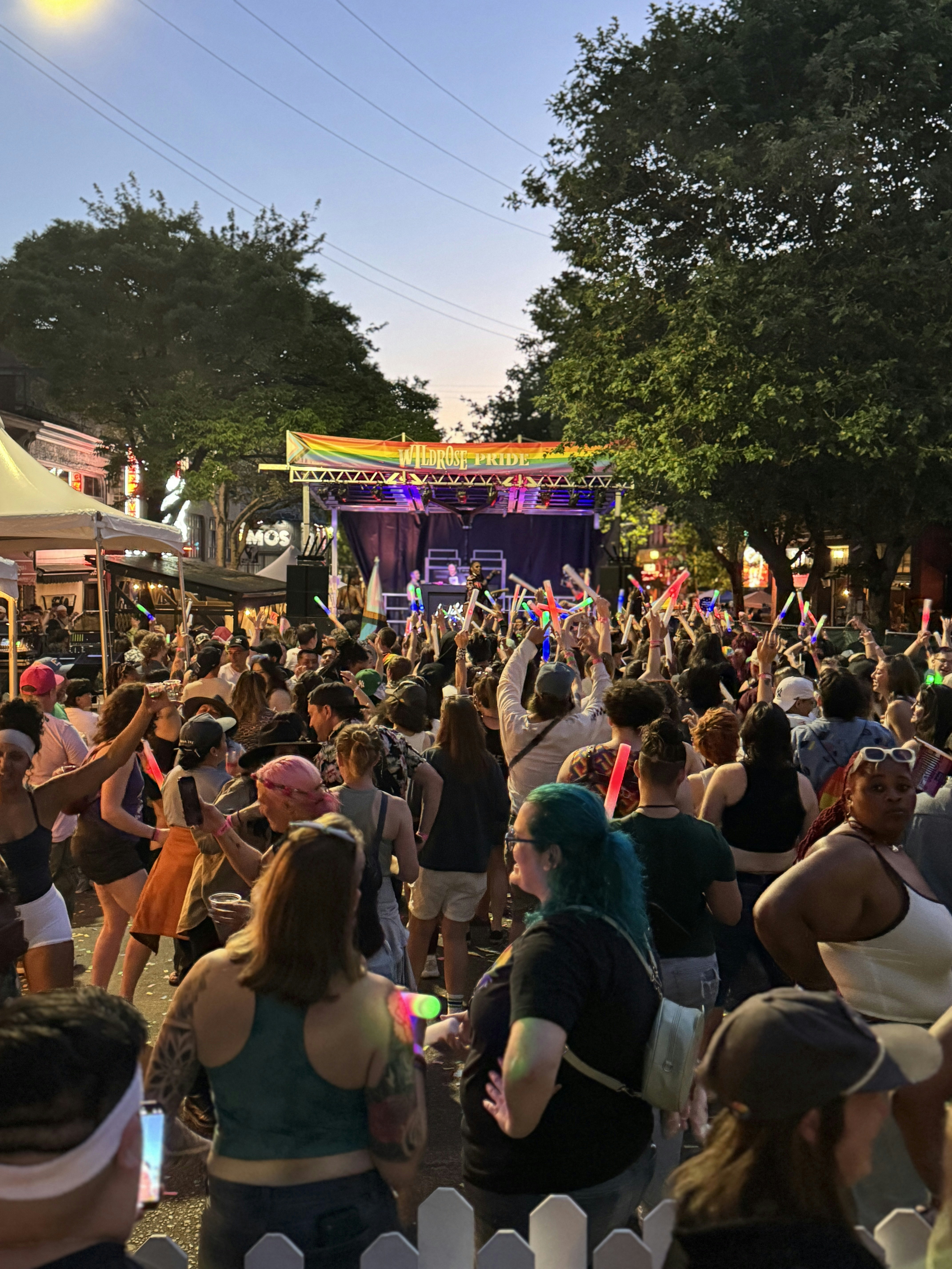 A crowd of people stand in front of a stage where a band plays; a rainbow-striped banner above the stage reads "Wildrose Pride."