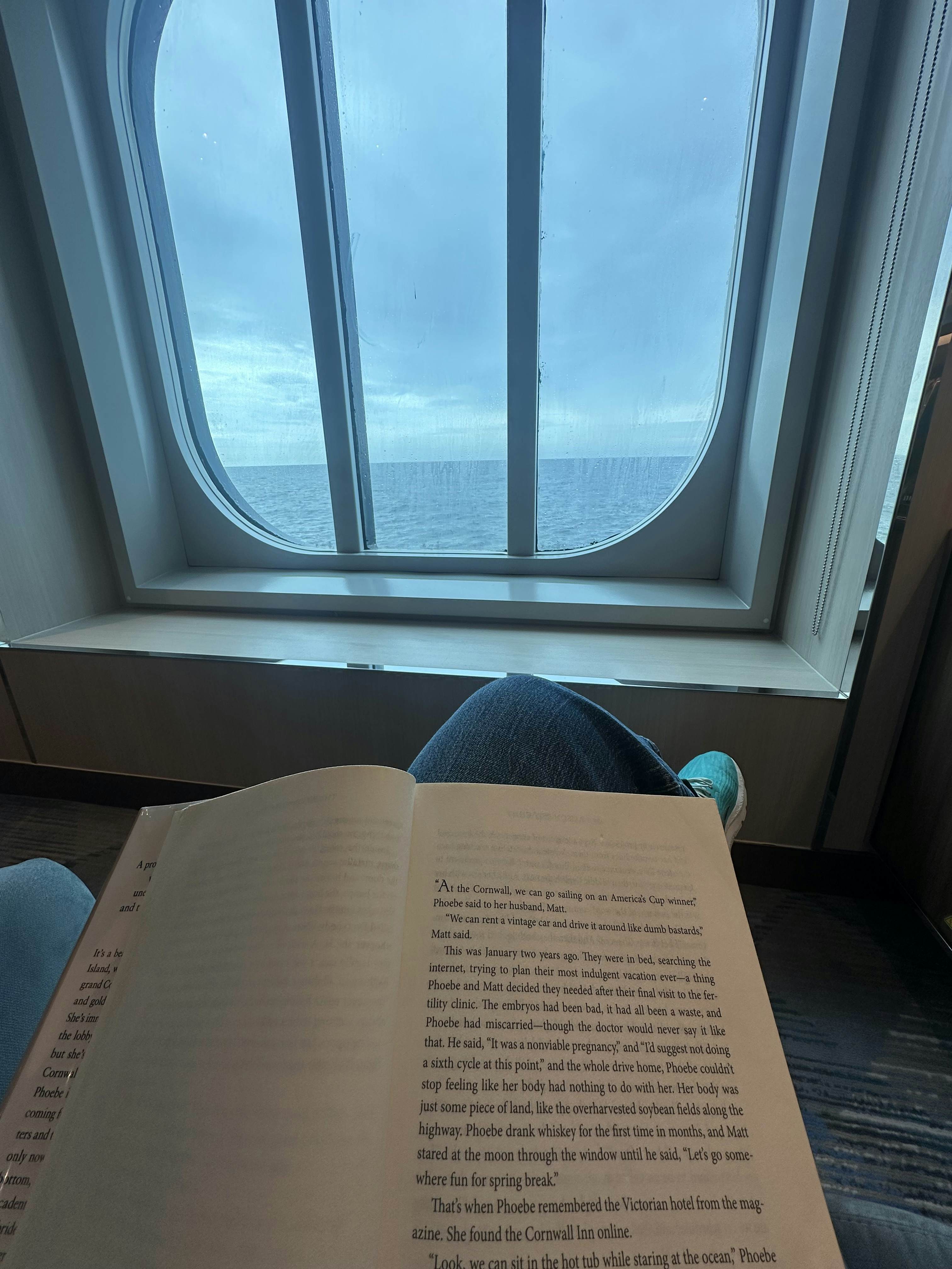 A notebook on a person's lap as they sit in a chair inside a cruise ship overlooking the ocean through a small window.
