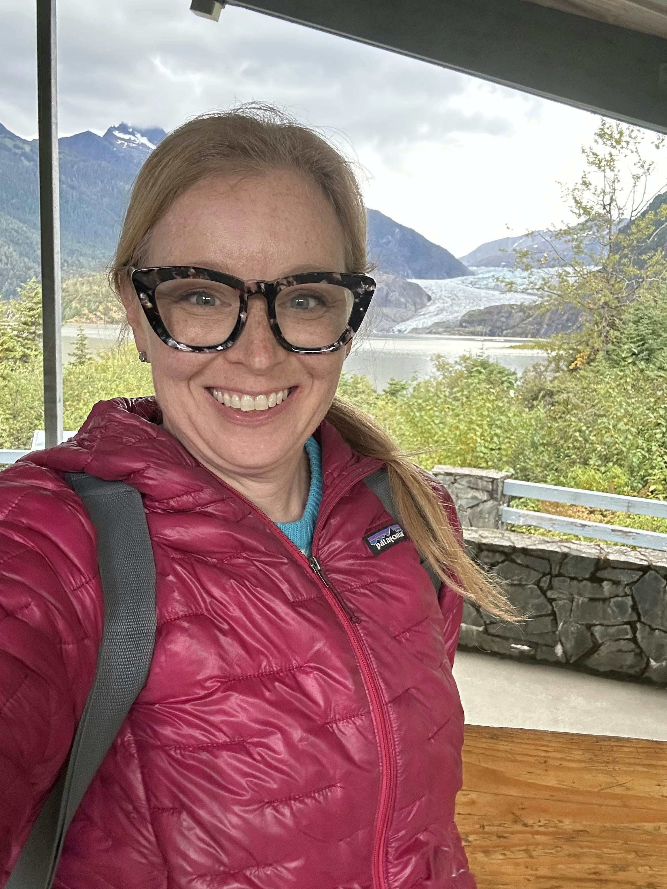 A woman wearing a red winter jacket and glasses smiles, with water and forest in the backbround.