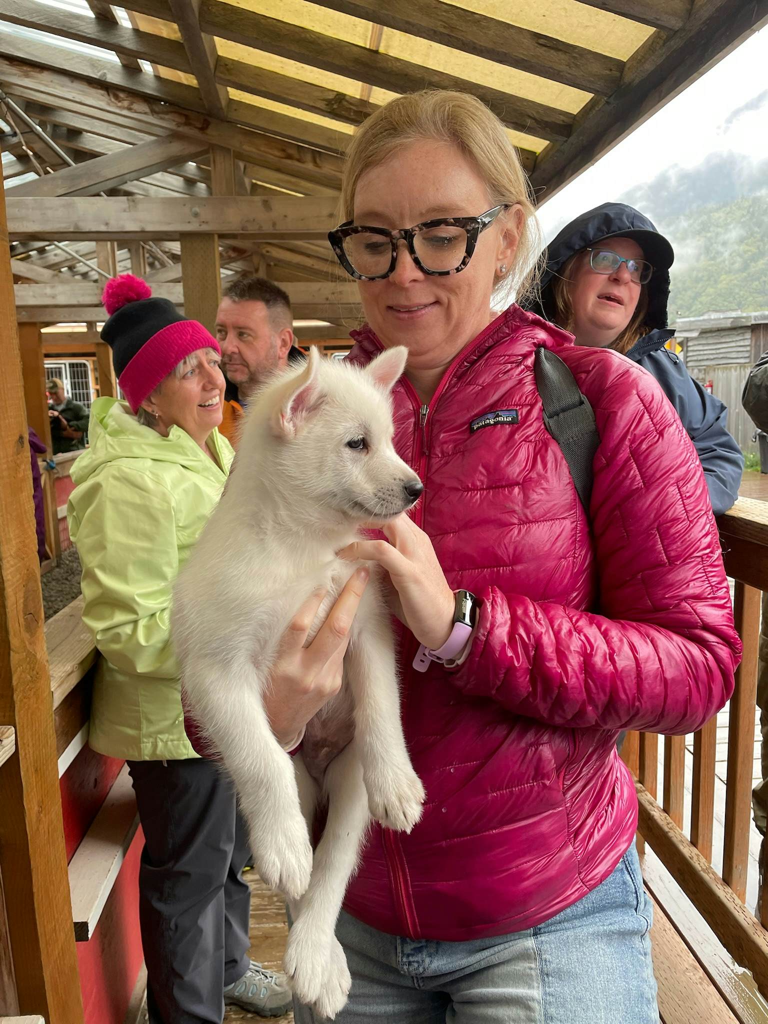 A woman wearing a red winter jacket and glasses holds a white, furry puppy.