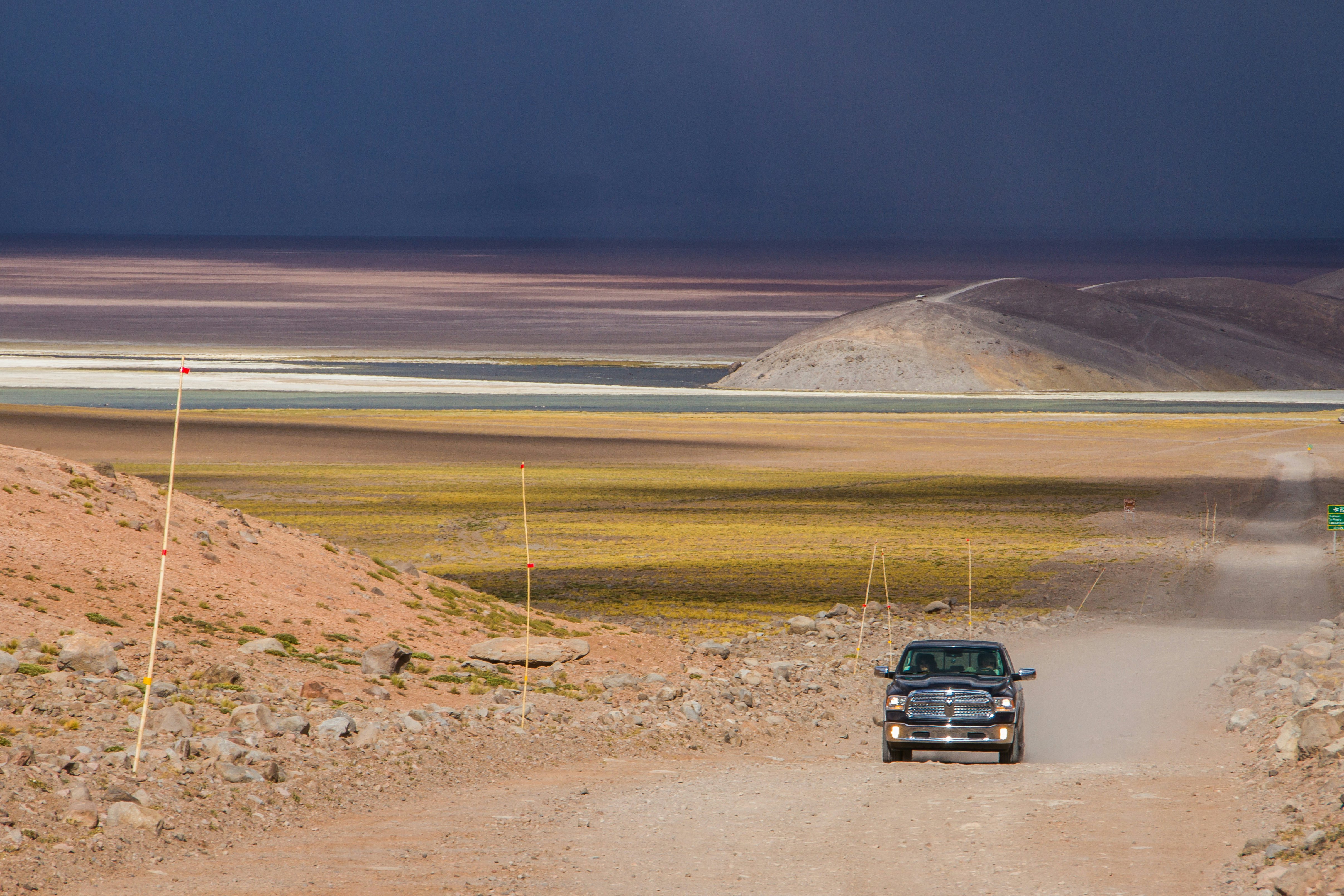A truck drives on a dirt road in a desert plain. The landscape in the distance forms bands of color.