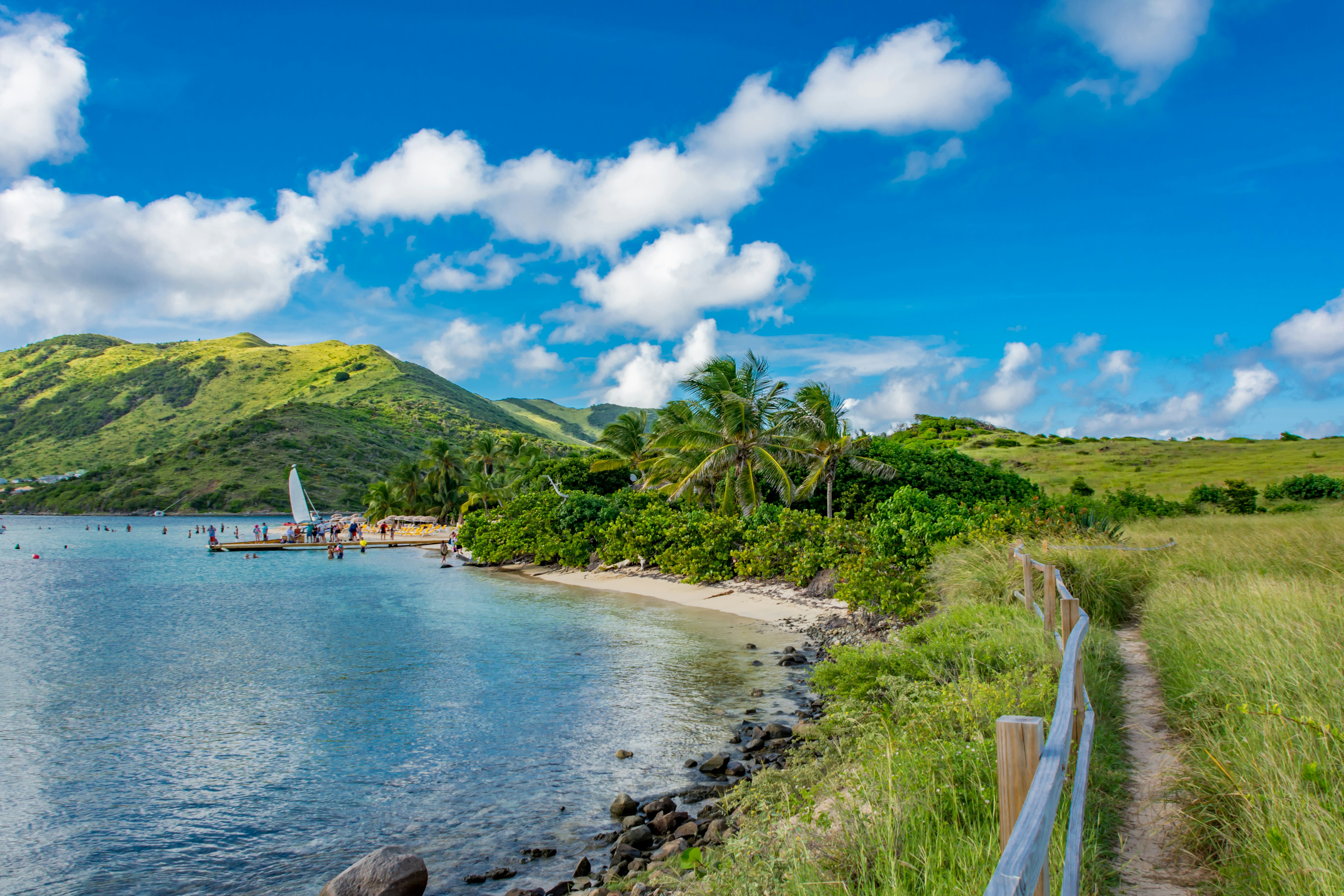 A bay with a sandy beach, people and low, forest-covered hills in the distance, and a narrow dirt pathway to the right on a sunny day.