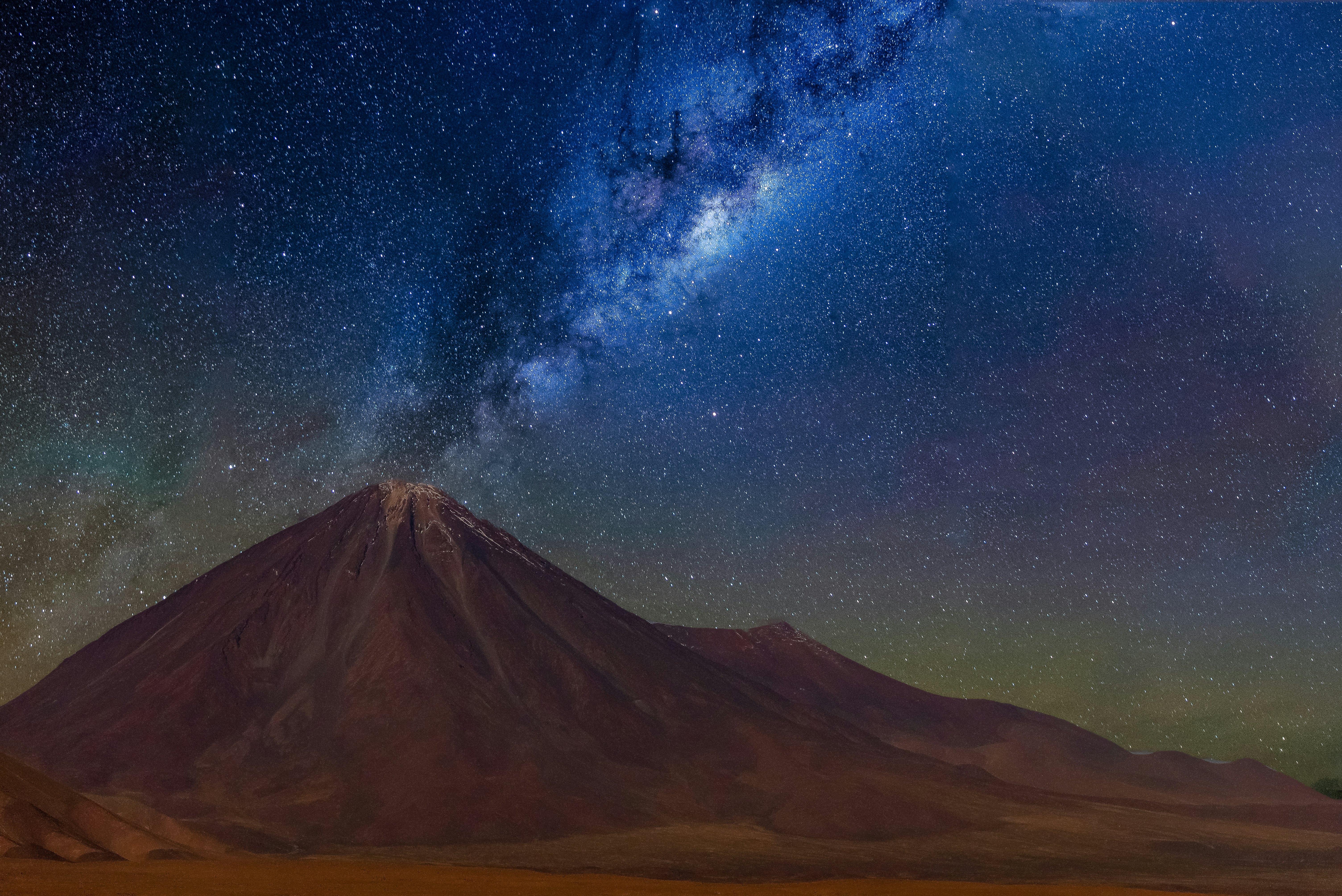 The Milky Way and thousands of stars are seen behind a volcano in a desert.