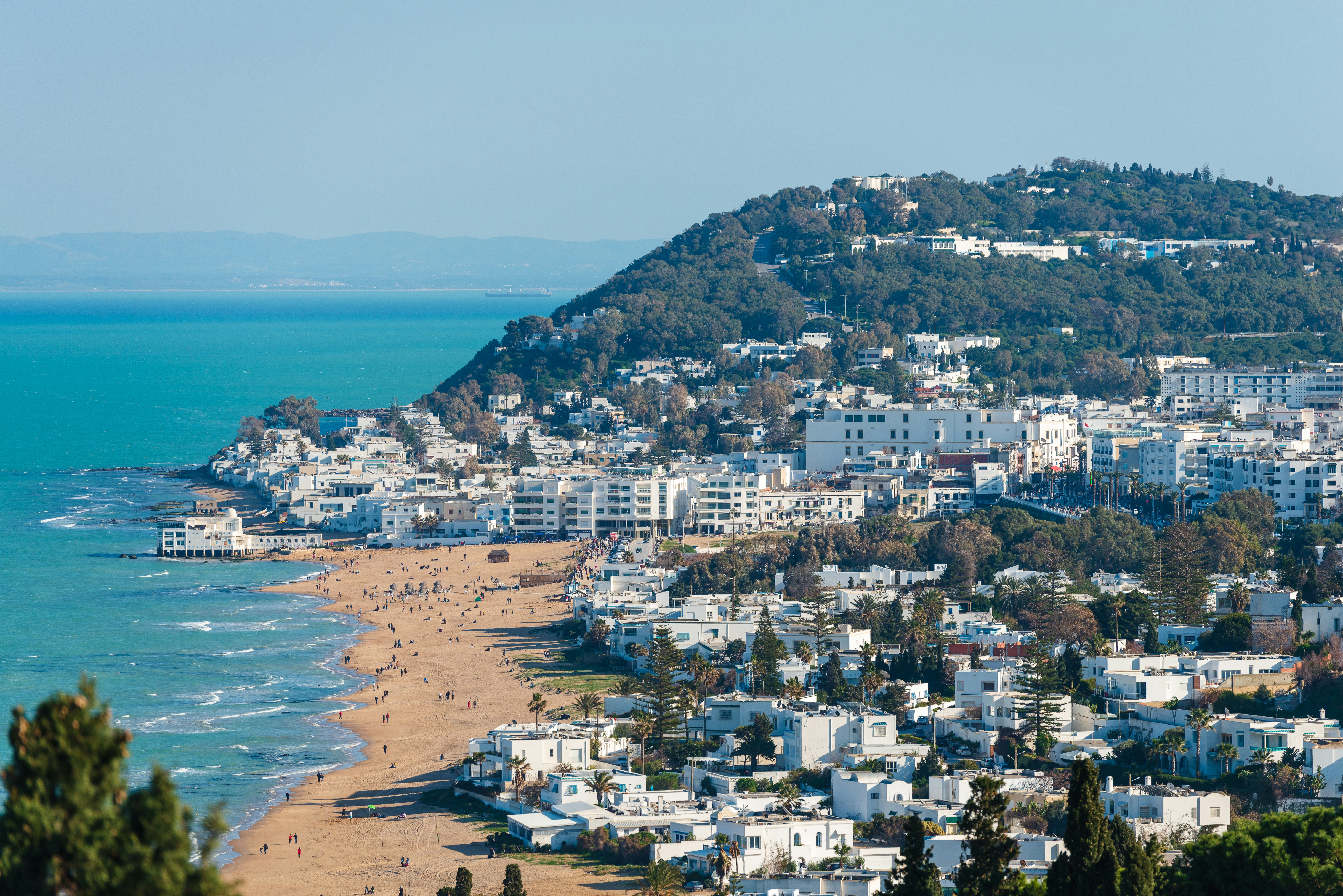 People on a city beach backed by white low-rise buildings covering a nearby hillside.