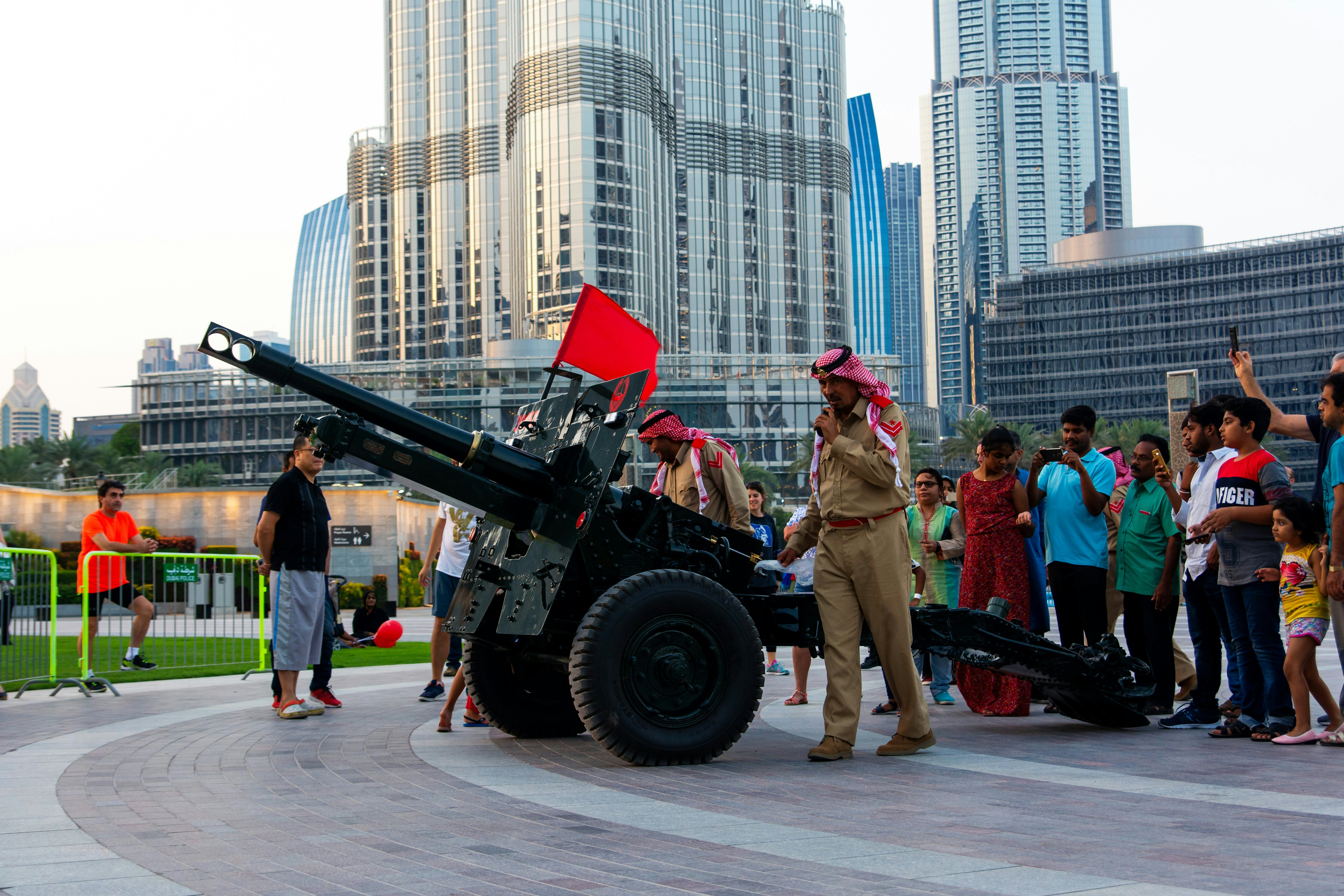 People stand near an artillery cannon in a city. Troops man the cannon, and skyscrapers are seen behind.