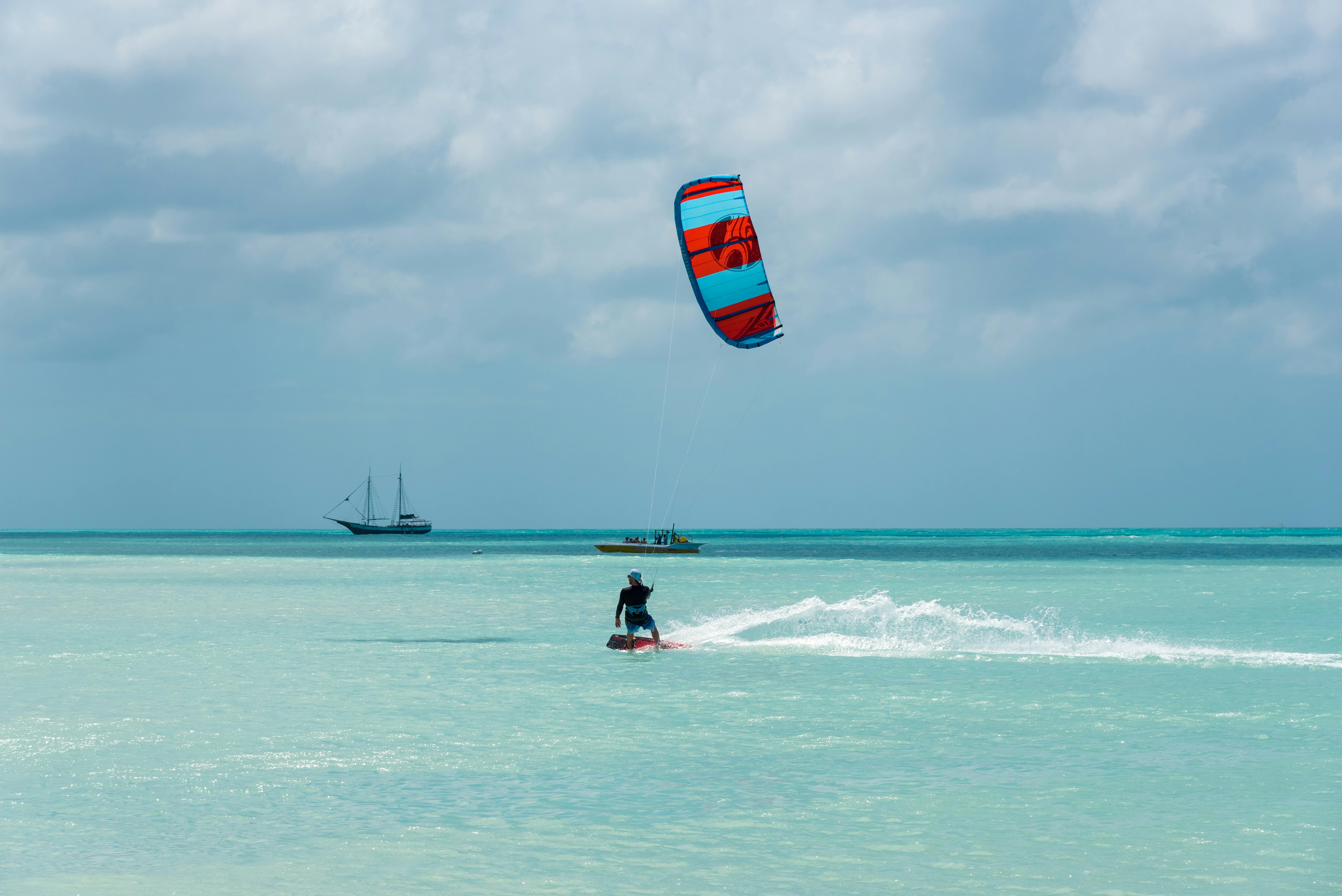 A person on a kiteboard in light blue water with a power boat and sailbot in the distance on a cloudy day.