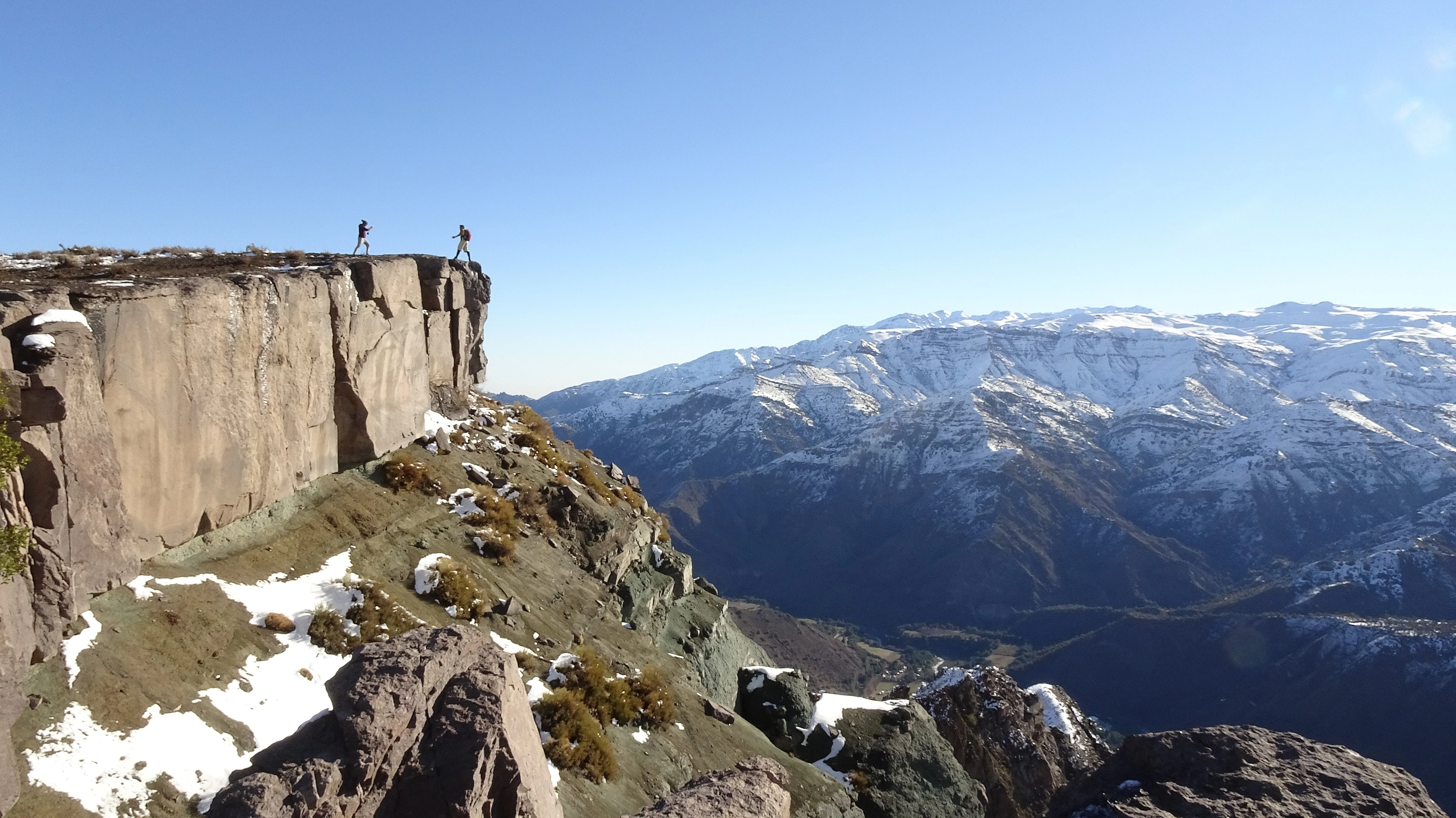 A wide shot of two people standing at the edge of a cliff overlooking a dramatic canyon with snowy mountains in the distance.