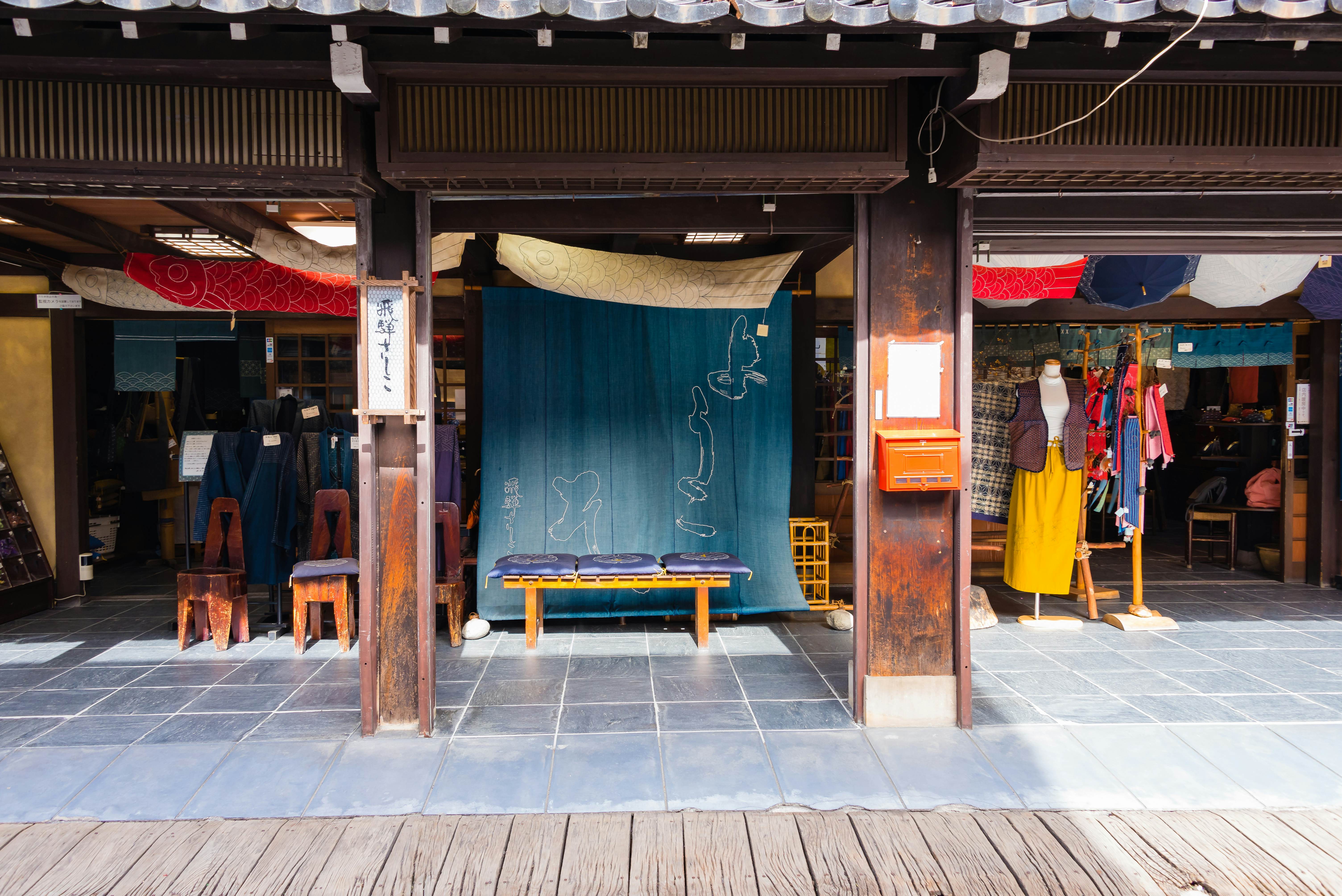 Exterior of fabric shop with a large indigo-dyed cloth in the center and a mannequin with long yellow skirt and brown vest to the right.