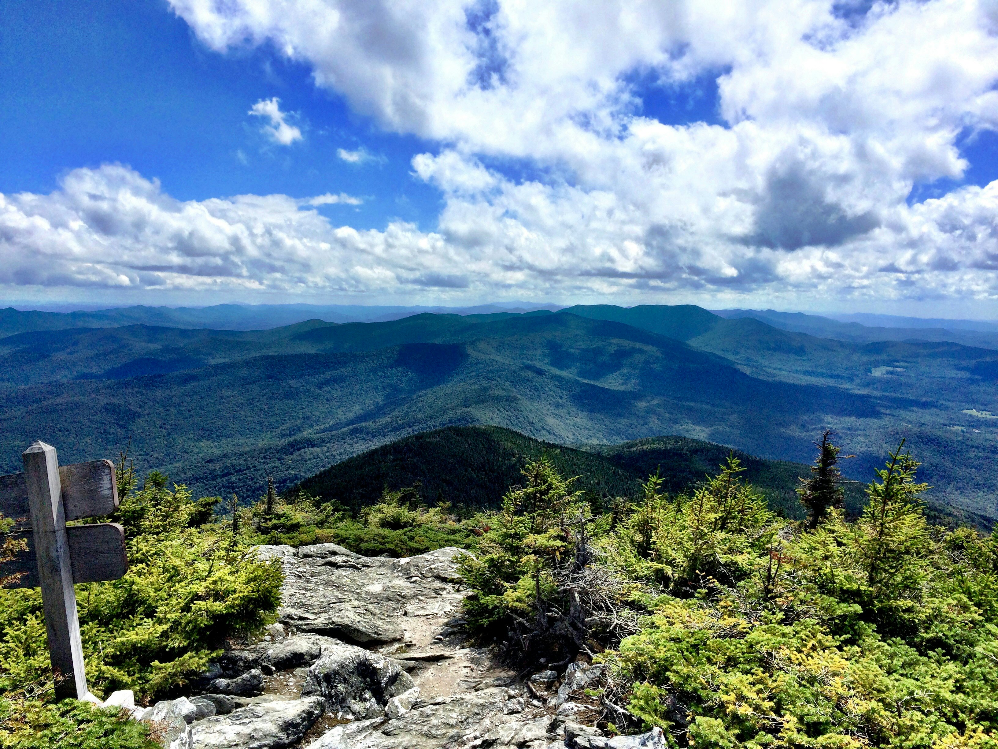 Low, rolling, forested mountains and wispy clouds as seen from a rocky mountain summit.