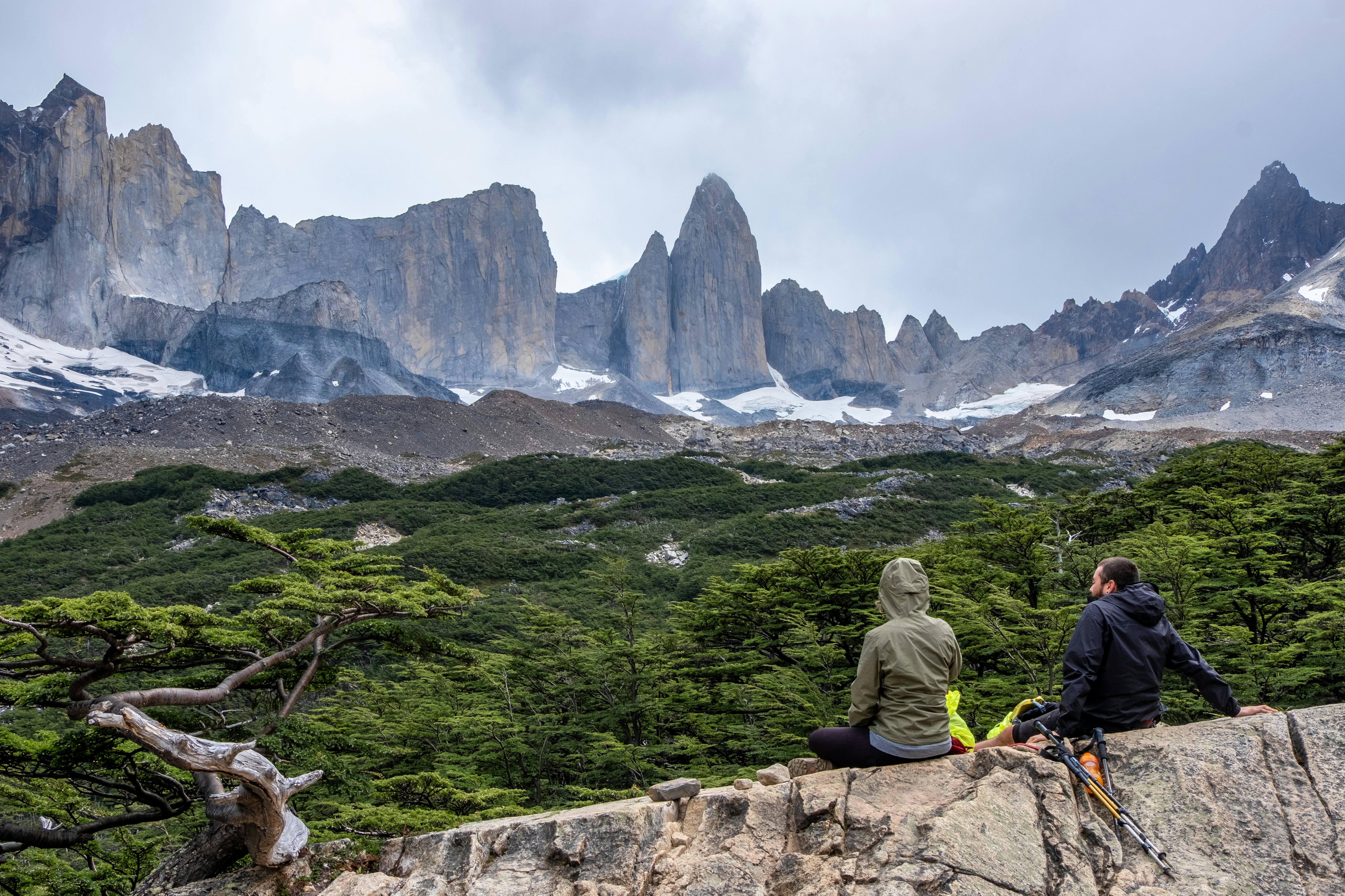Two hikers sit on a rock looking at craggy mountains across a valley.