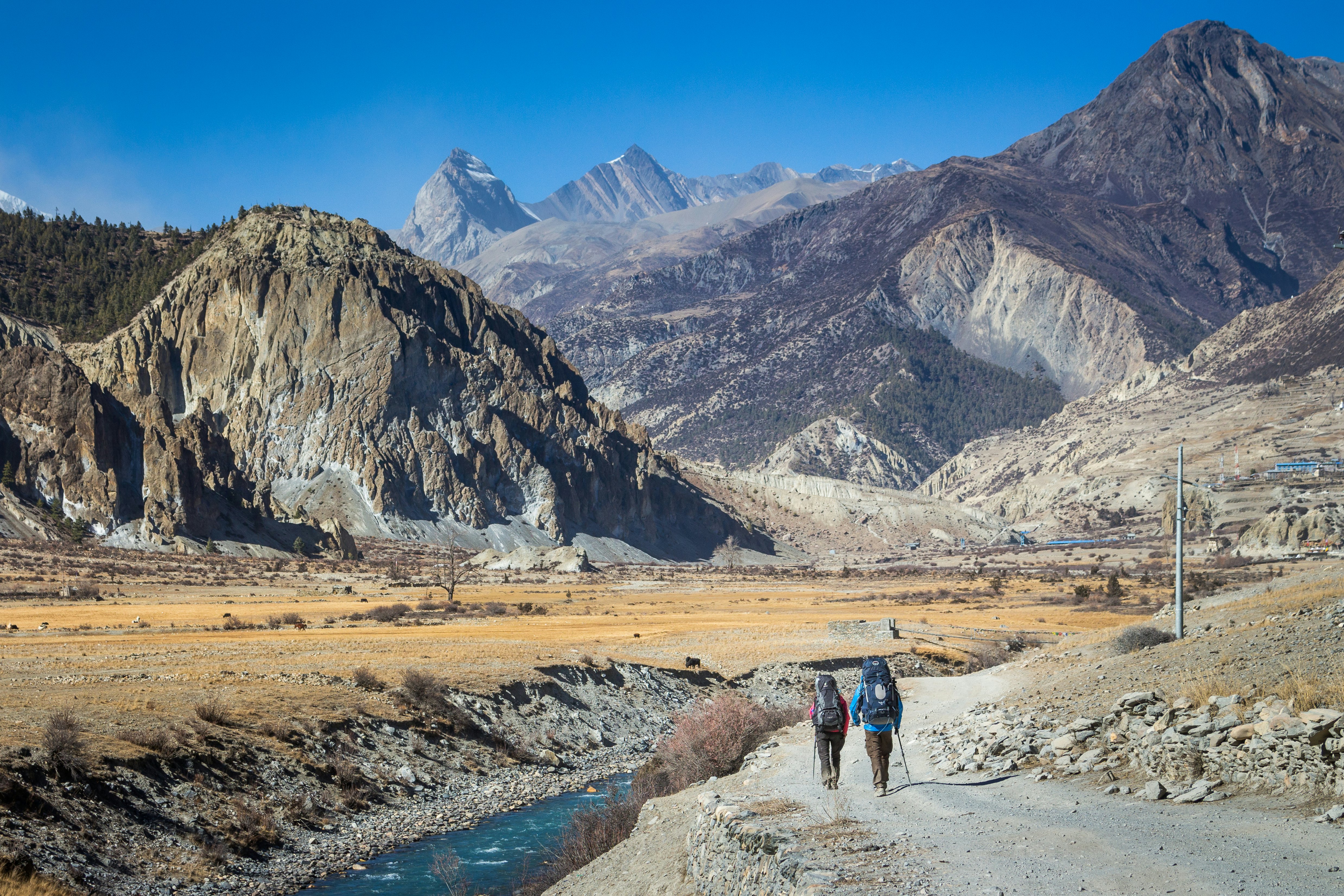 Two trekkers on a path following a small river in a valley surrounded by rocky slopes and high peaks in the distance.