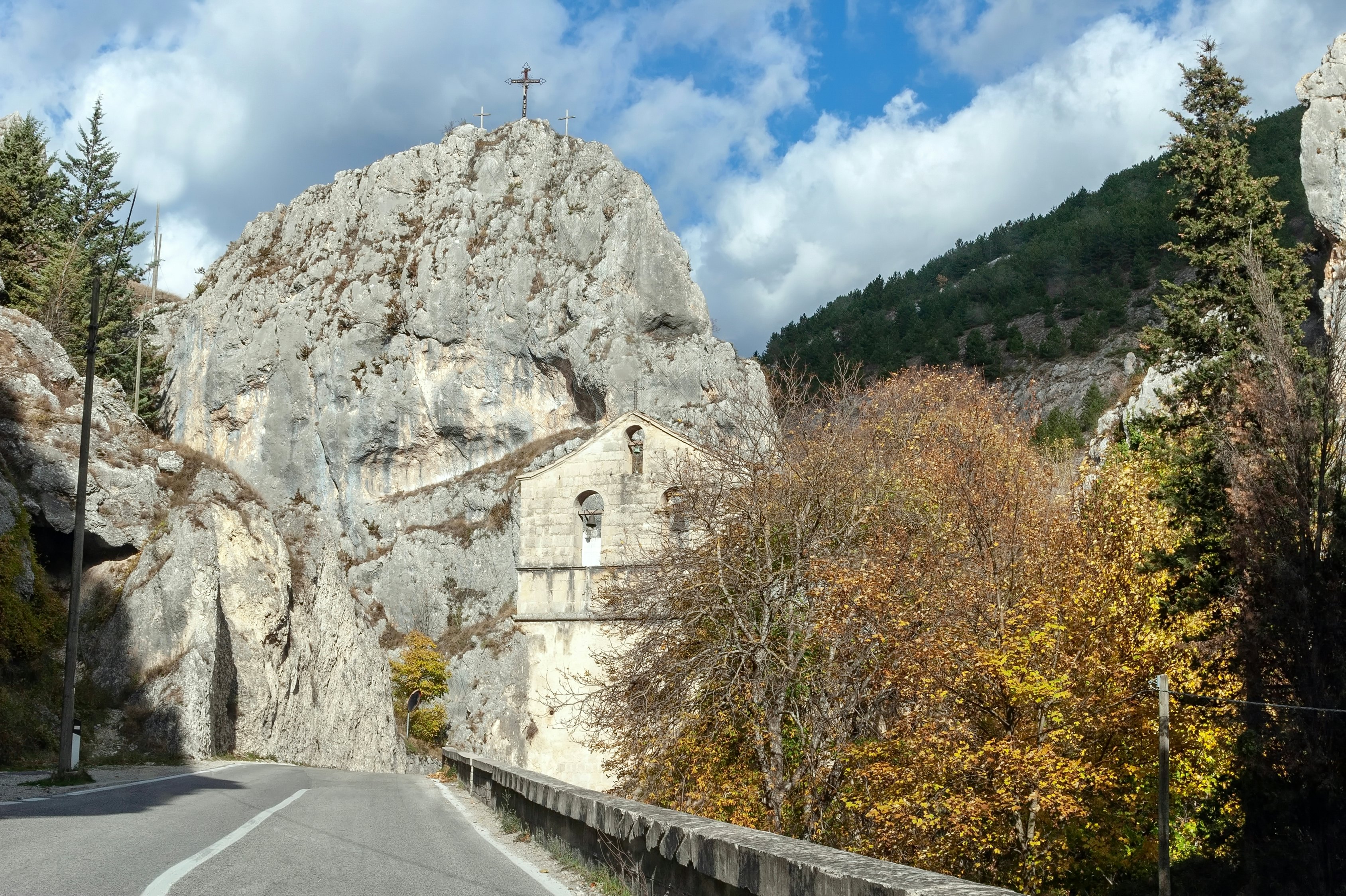 A road leads through a mountain pass, by a stone church. A rocky outcrop topped by a cross is seen in the distance.