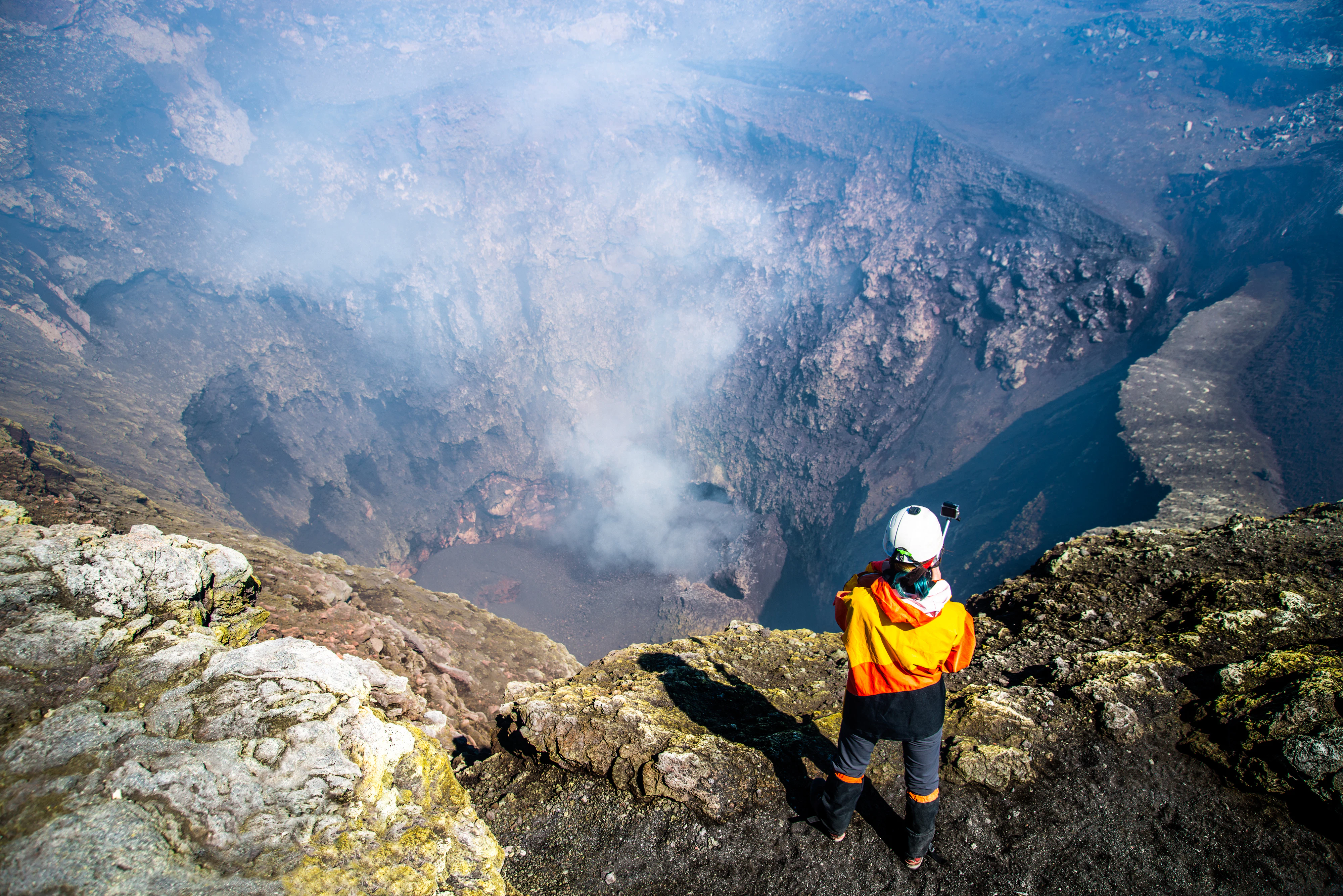 An overhead shot of a hiker in a helmet peering into the deep crater of a volcano.