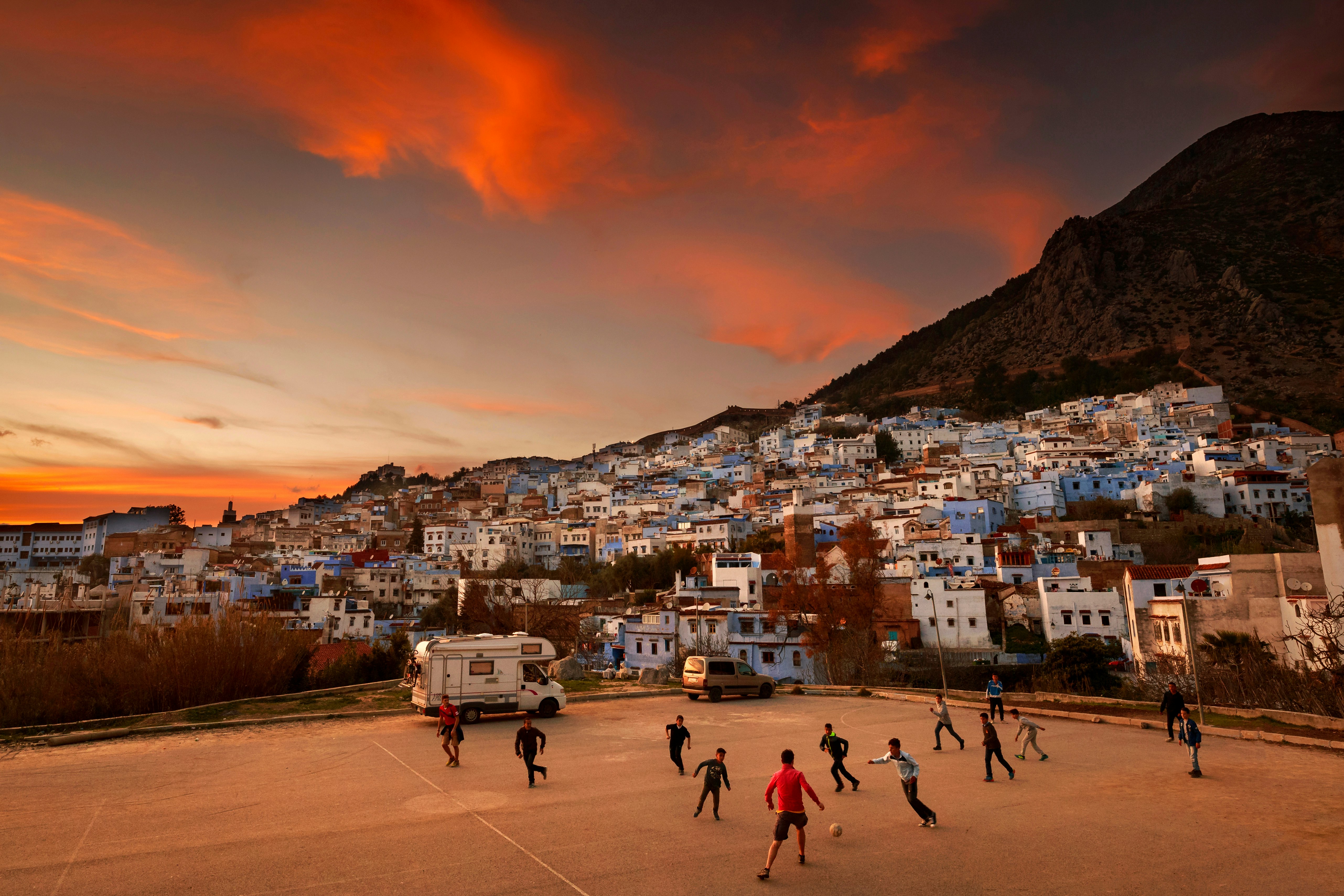 A group of teenagers plays football as the sun sets over a hillside village with houses painted in different shaddes of blue.