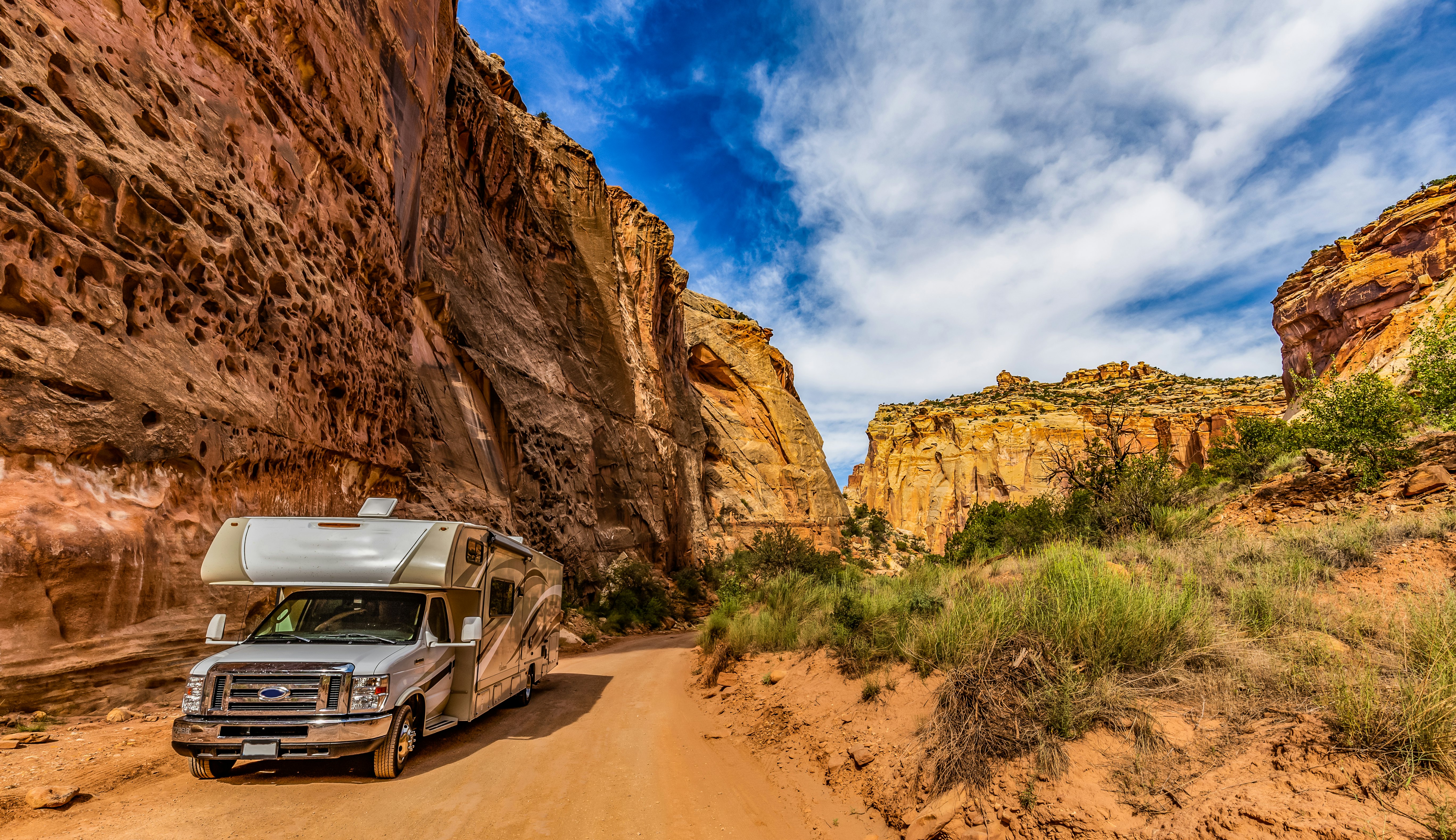 A camper van on a dusty trail along a sheer yellow and red rock face.