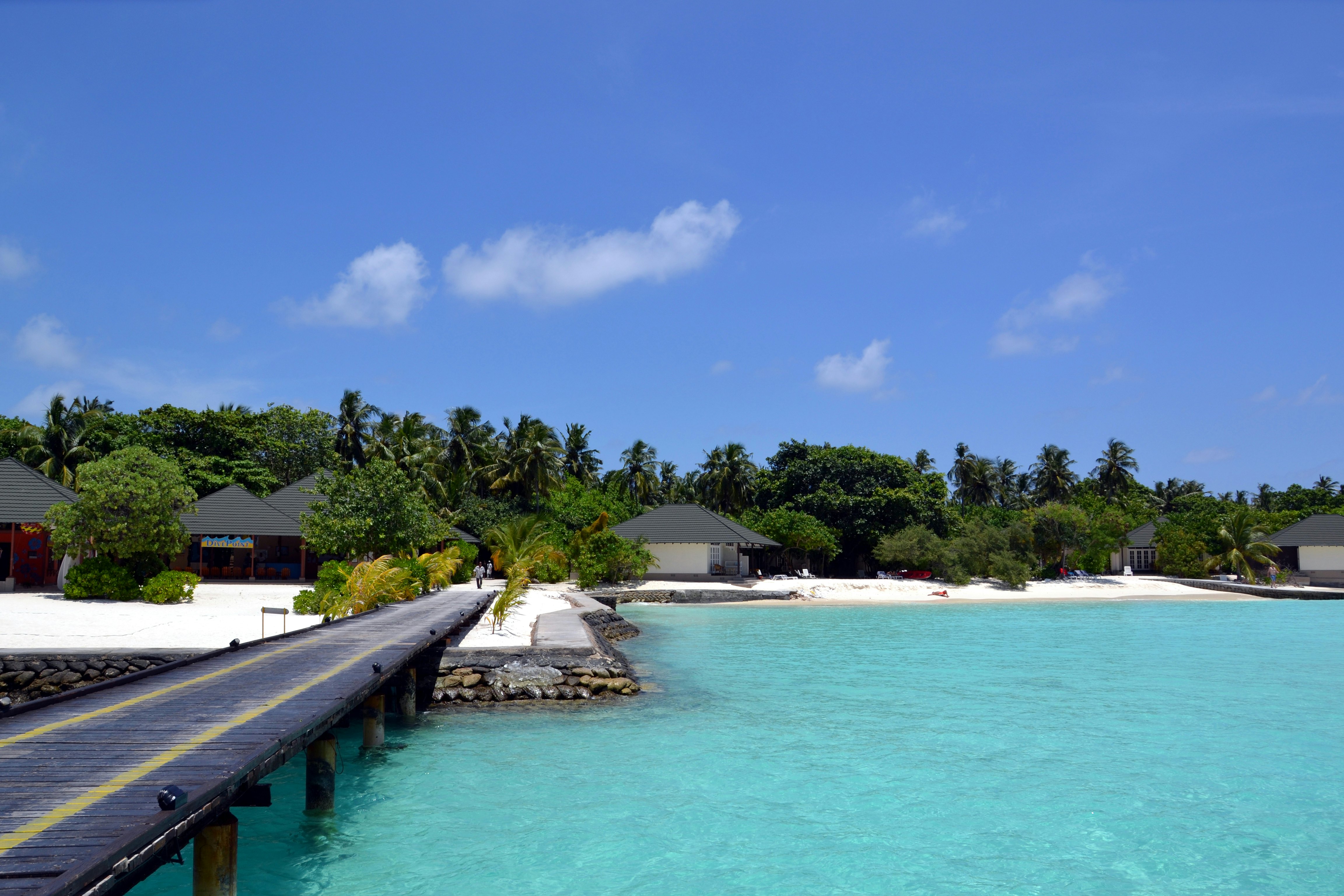 A dock over clear blue waters. Small building with sloped roofs dot the shore