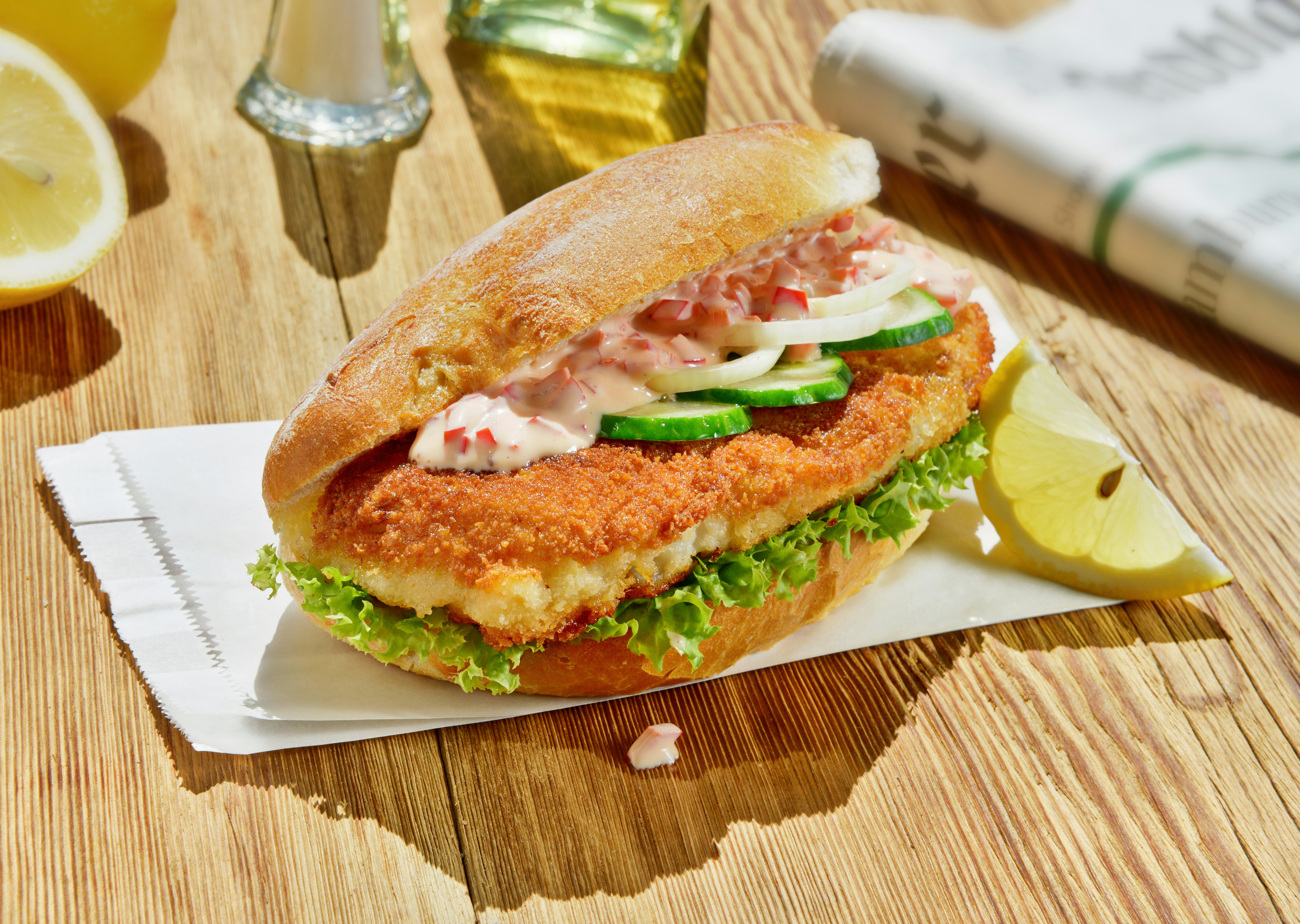Fried fish on a long crusty roll with lettuce, cucumber and onions sitting on top of a small, white takeout bag on a wooden table. A lemon slice leans agains the sandwich on a sunny day.