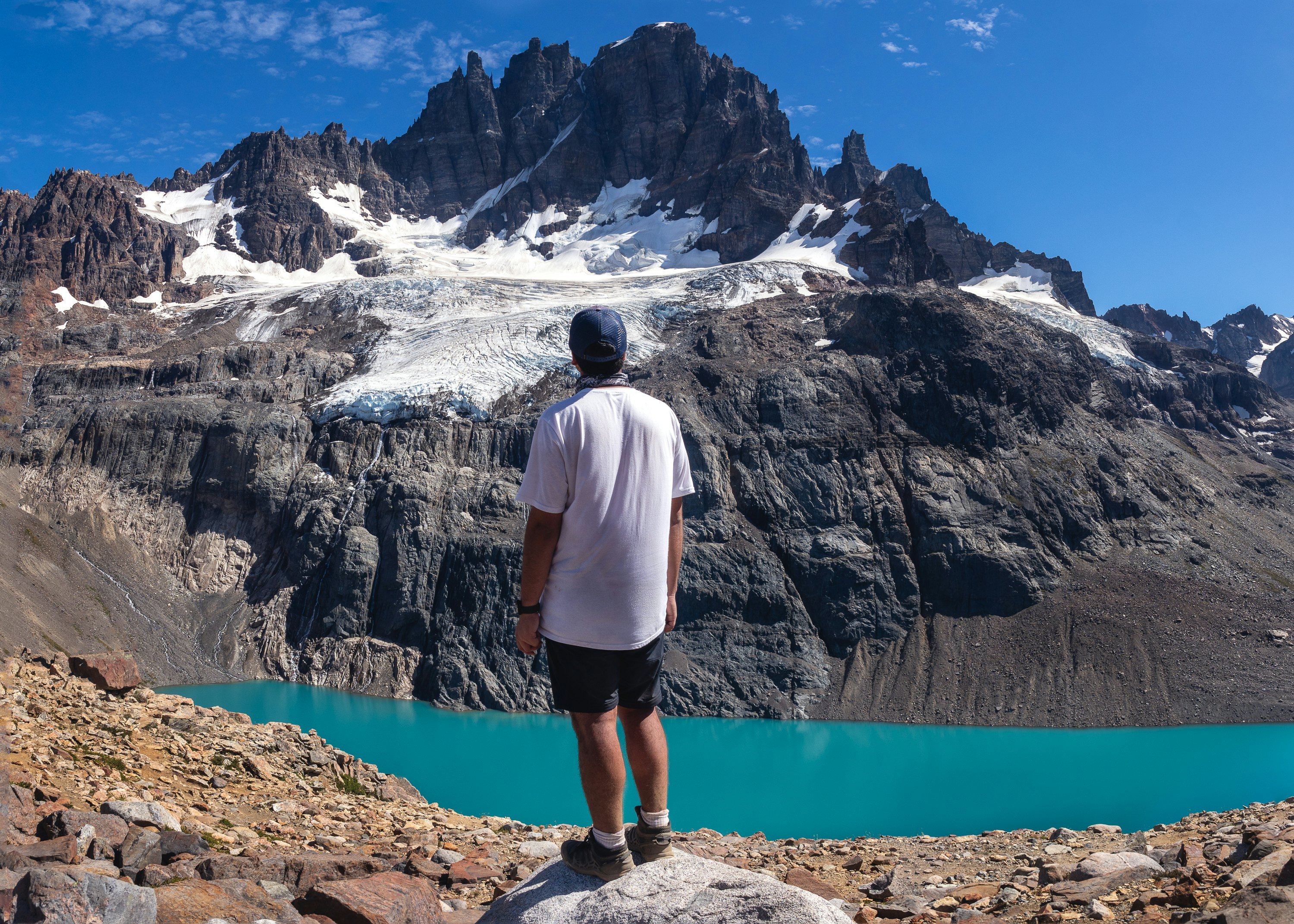 A man in a white t-shirt is seen from behind standing in front of a glacial lake looking at a huge, snow-covered mountain.