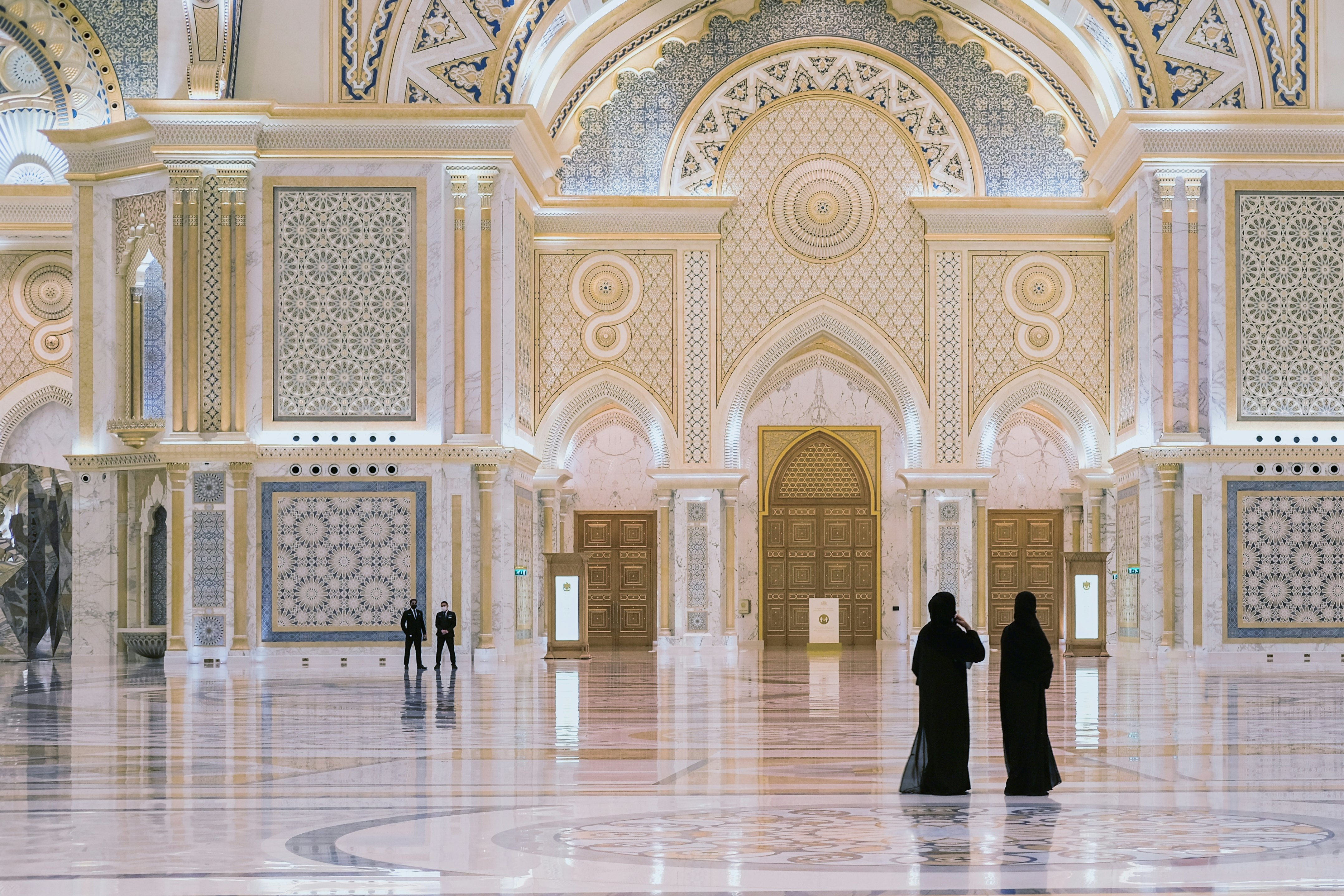 Two women wearing dark clothes covering their heads down to their feet in a grand palace room with detailed tiling all over the walls and floor.
