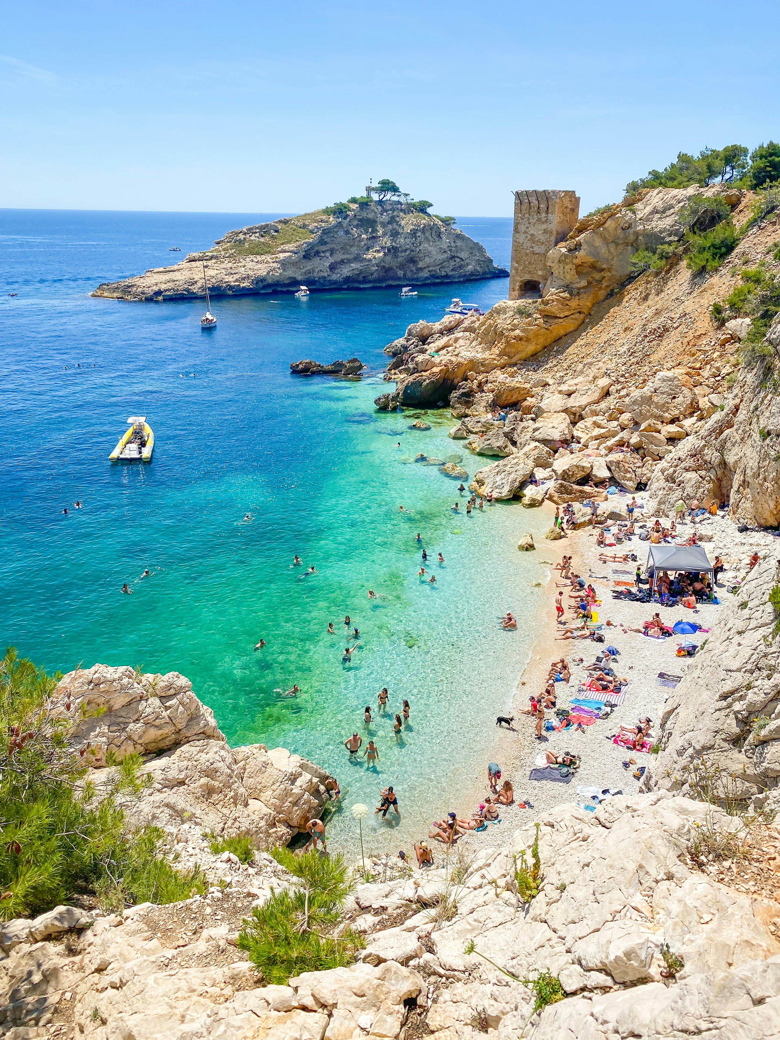 Sunbathers on a narrow stretch of beach backed by cliffs. People swim and splash in the turquoise ocean.
