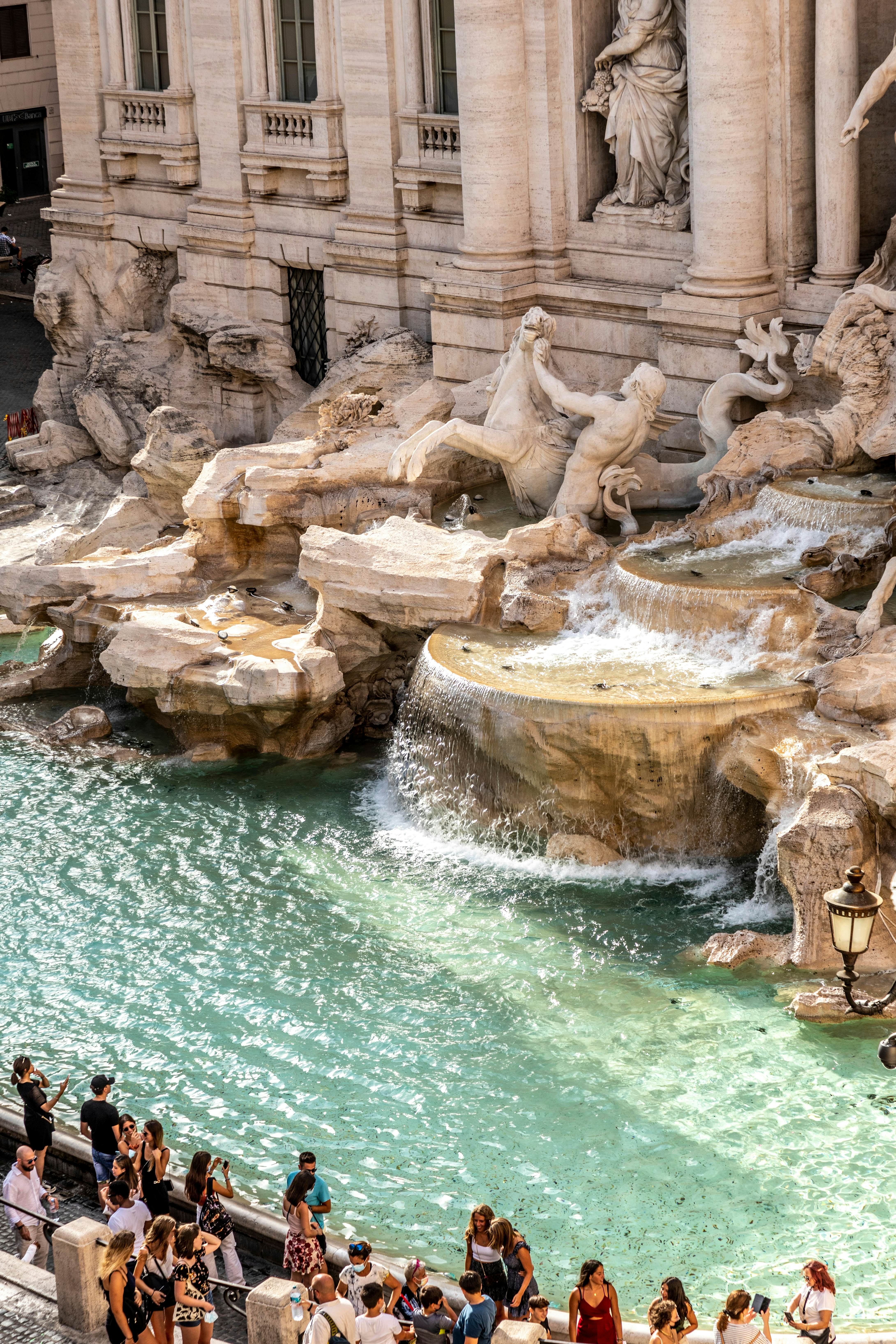 People gather in front of the clear waters of the Trevi fountain, right in front of the giant marble statues.