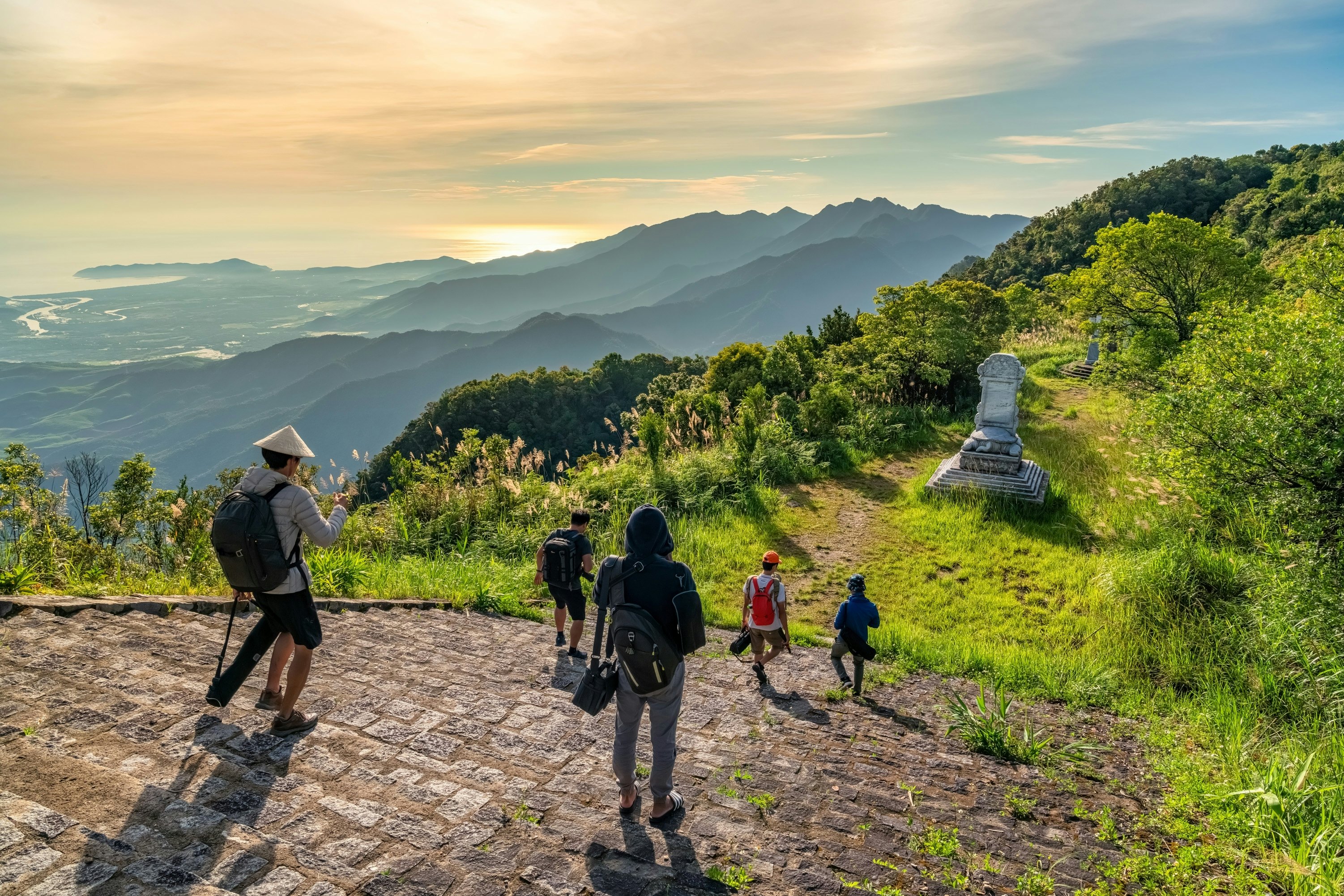 People walk down steps at the top of a hill with views in the distance of the sunrise over the sea.