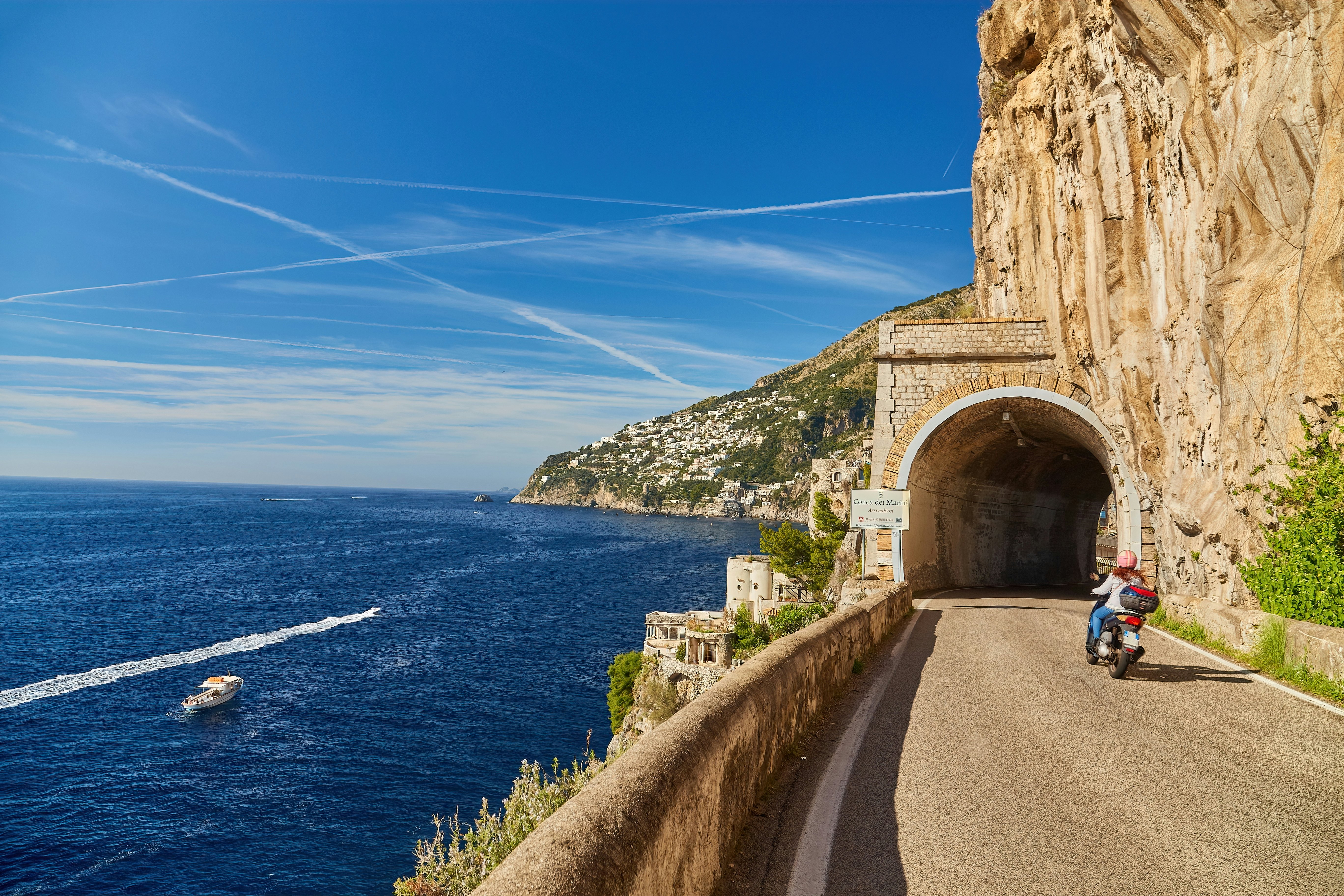 A scooter approaches a tunnel on a road hugging the cliffs of a coast. Boats are seen in the sea below the road.