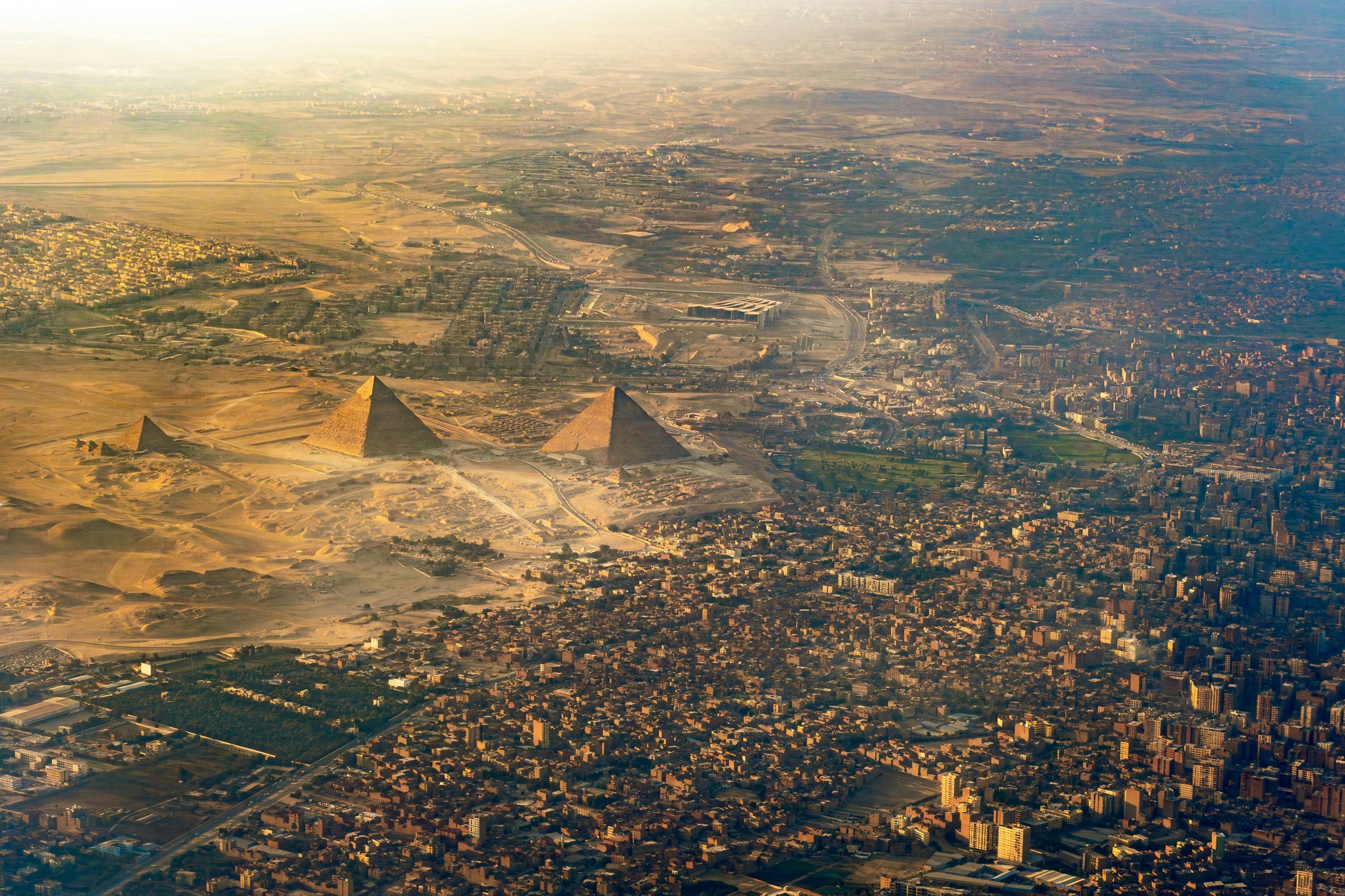A large sprawling city gives way to sandy desert. Three large pointed pyramids stand between the city and desert.