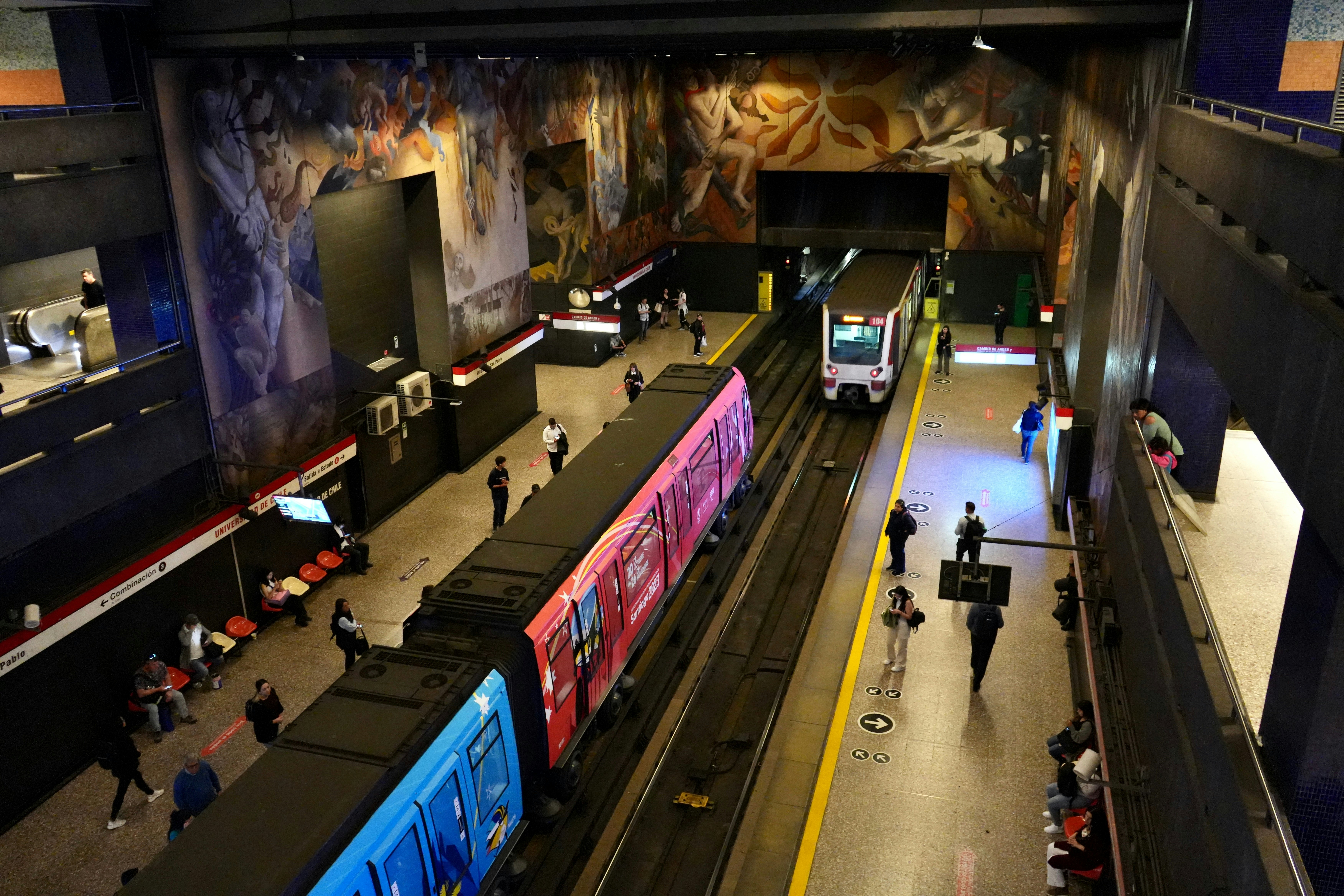 An overhead view of trains in an underground metro station and people on the platforms. Painted murals cover the station’s walls.