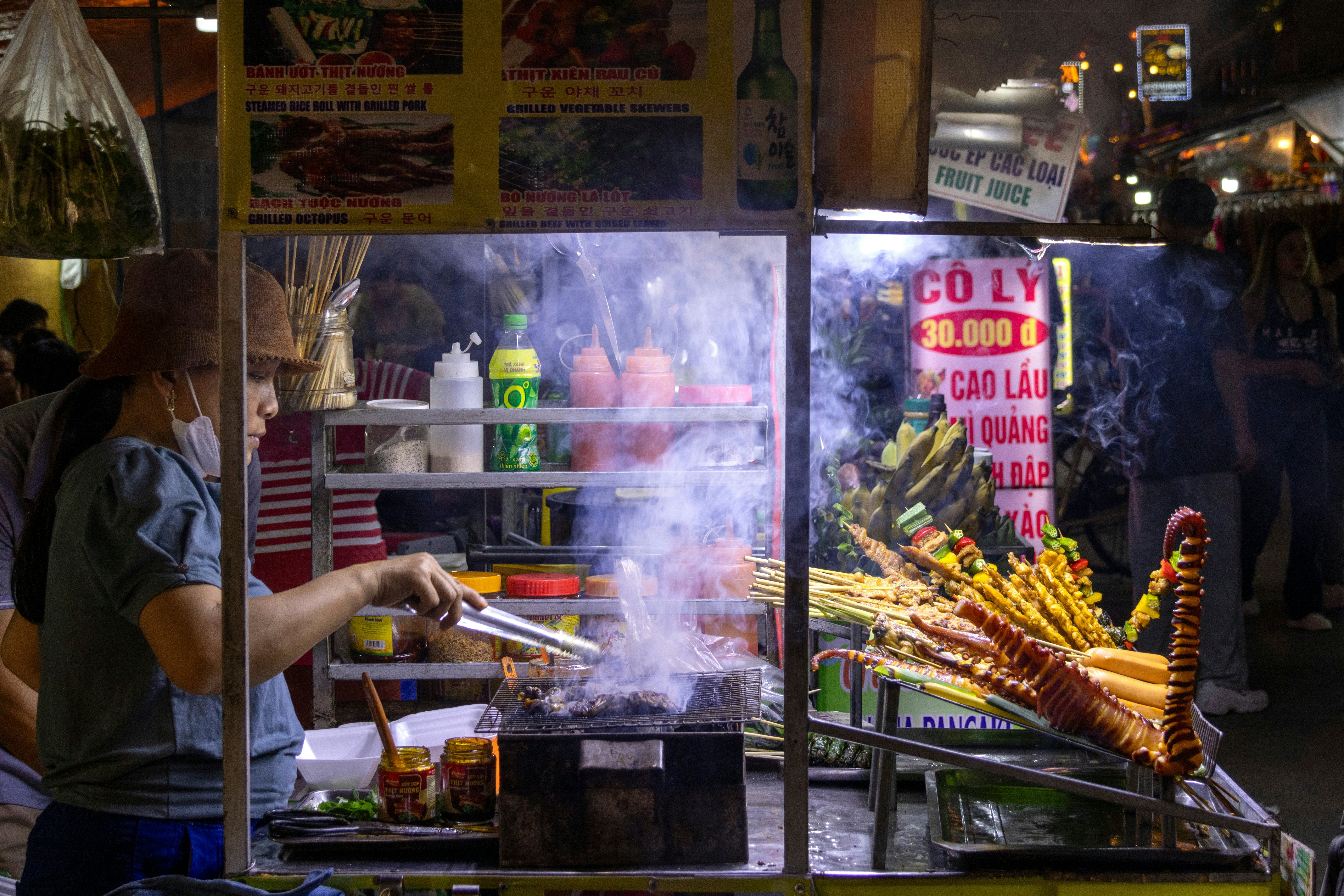 A woman grills meat over a smoky fire at a night market.