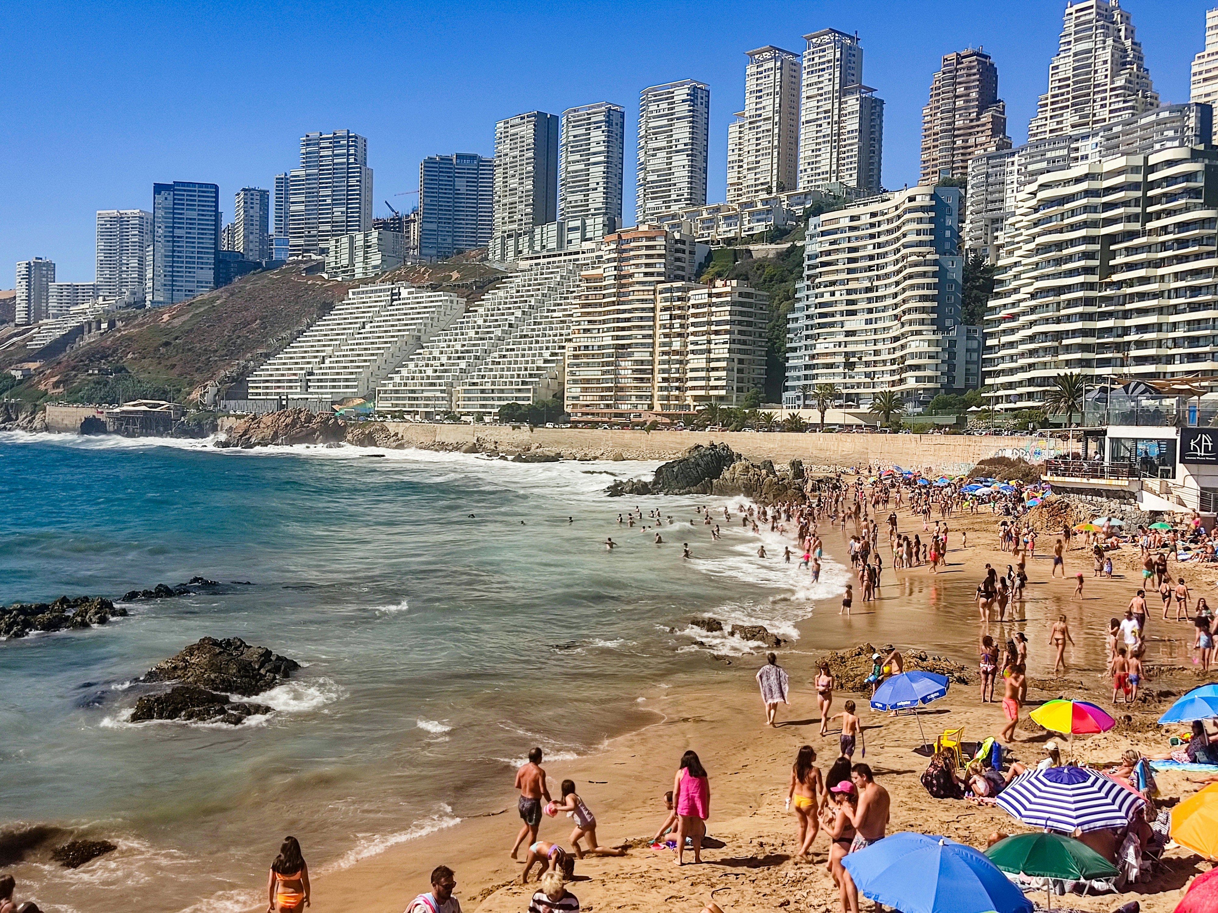 People are pictured on a crowded beach with apartment towers lining a bluff in the distance.