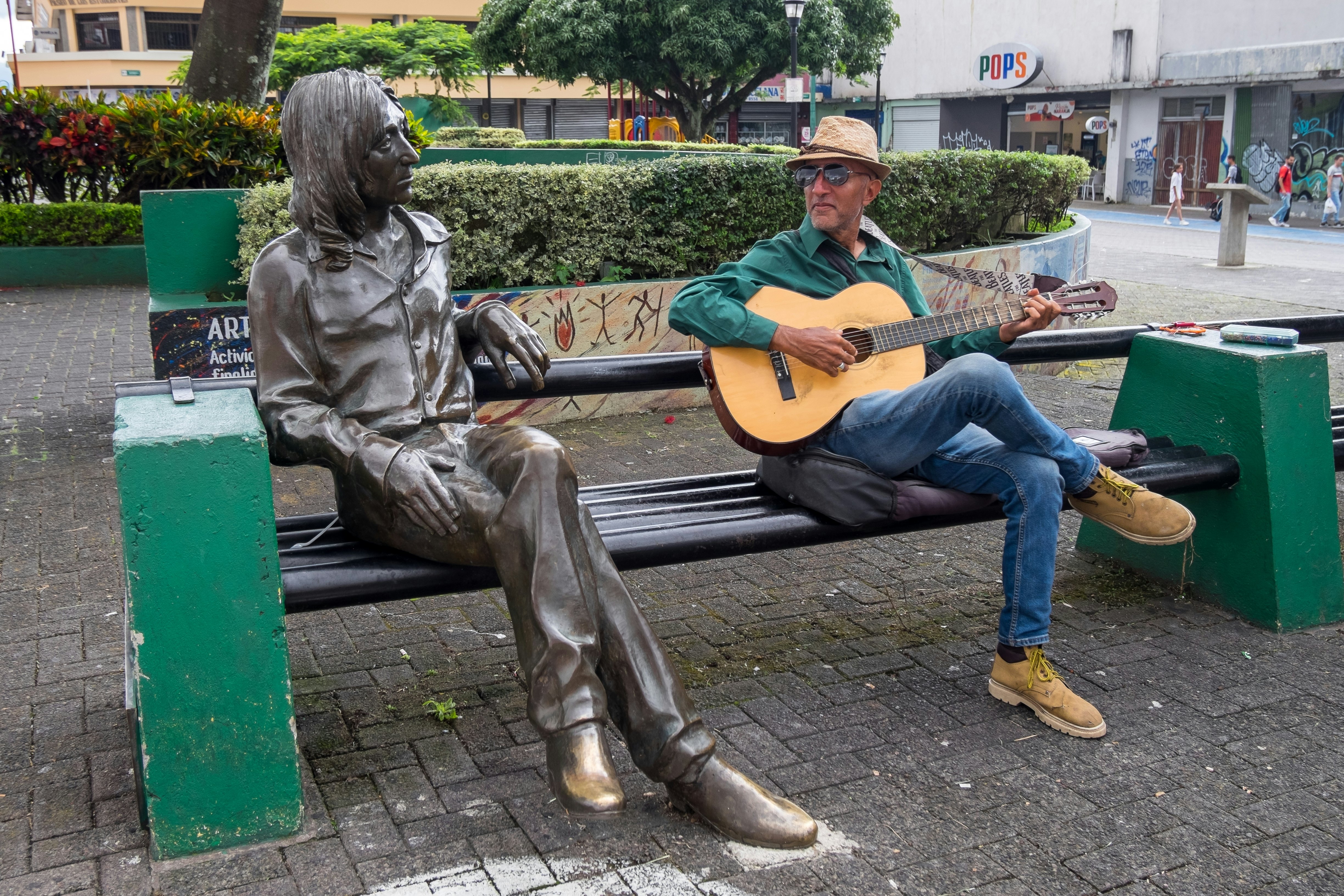 Sitting next to a life-size bronze statue, a man plays a guitar on a park bench in a city.