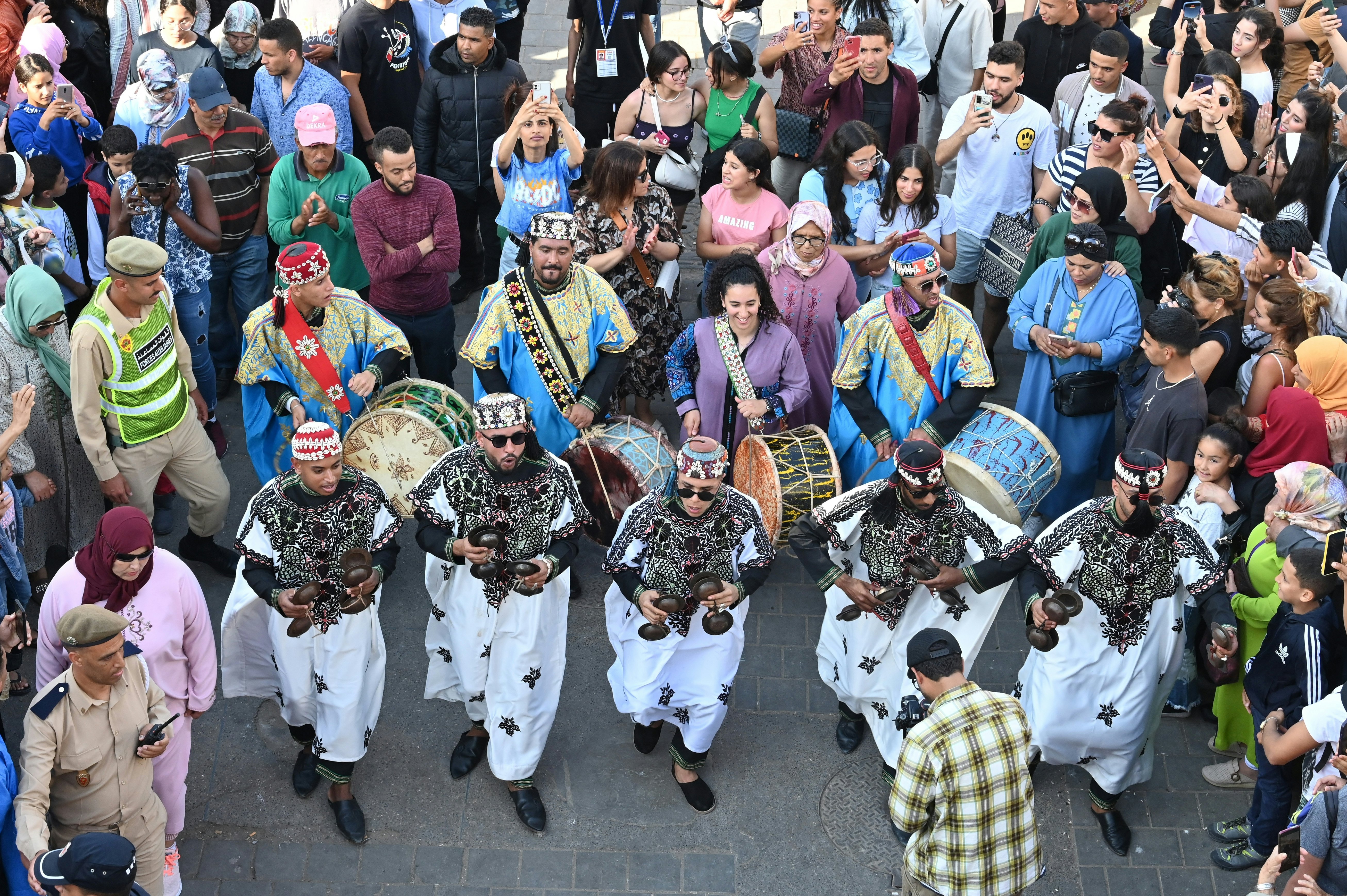 A parade of people and drummers march down the street surrounded by a crowd of onlookers.